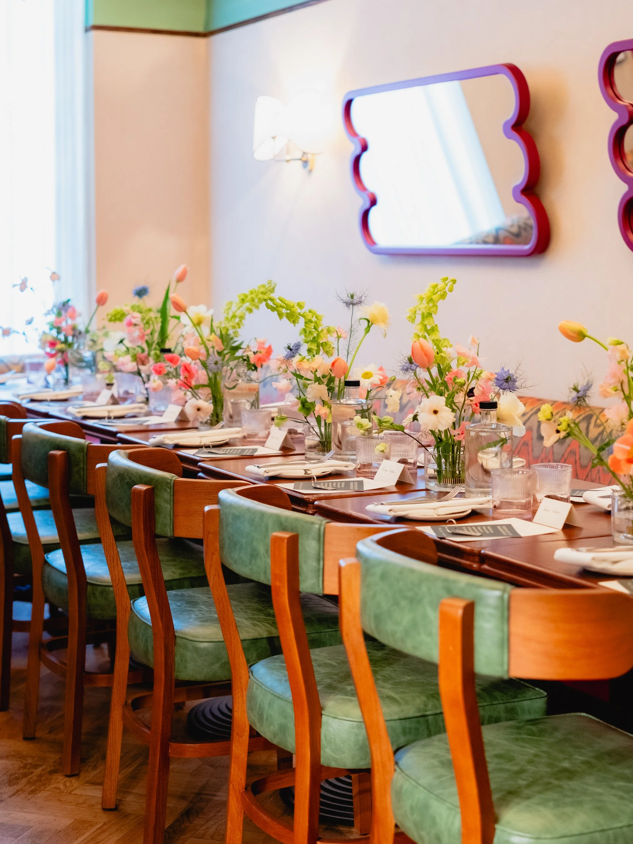 A long dining table decorated with pink, white, and yellow flowers in glass vases, with place settings, napkins, and menus underneath the flowers. The table is surrounded by wooden chairs with green cushions. There are colorful mirrors and a sconce o