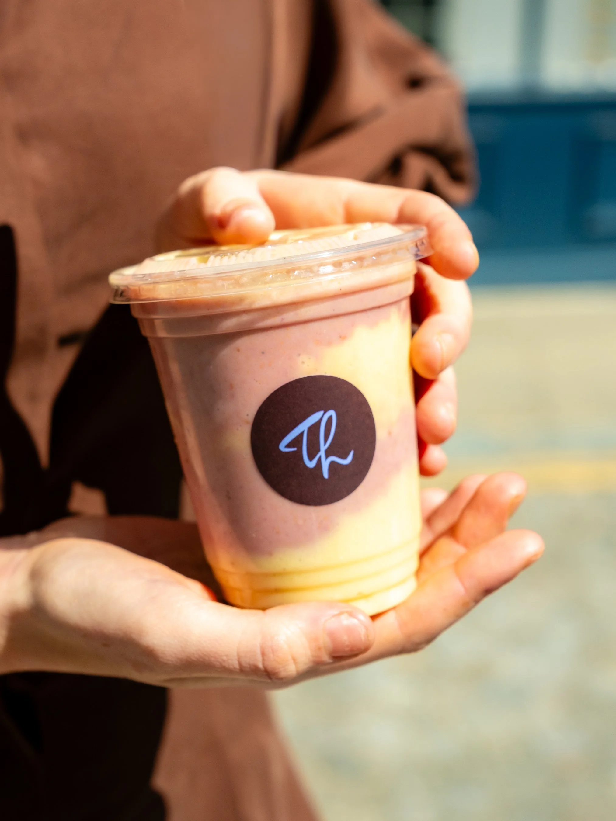 A person holding a cup of smoothie with a plastic lid, outdoors in natural light.