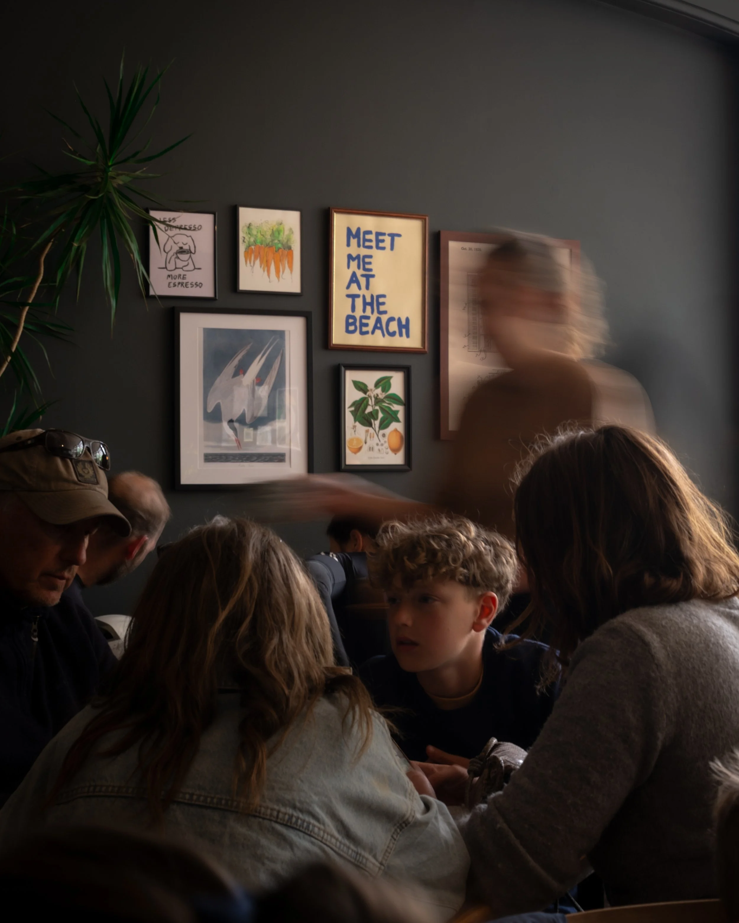 A group of people gathered indoors, with a dark grey wall on which various framed artworks and posters are hanging. One poster reads 'Meet Me at the Beach' in bold blue letters. There is a green plant with long, spiky leaves on the left side of the i