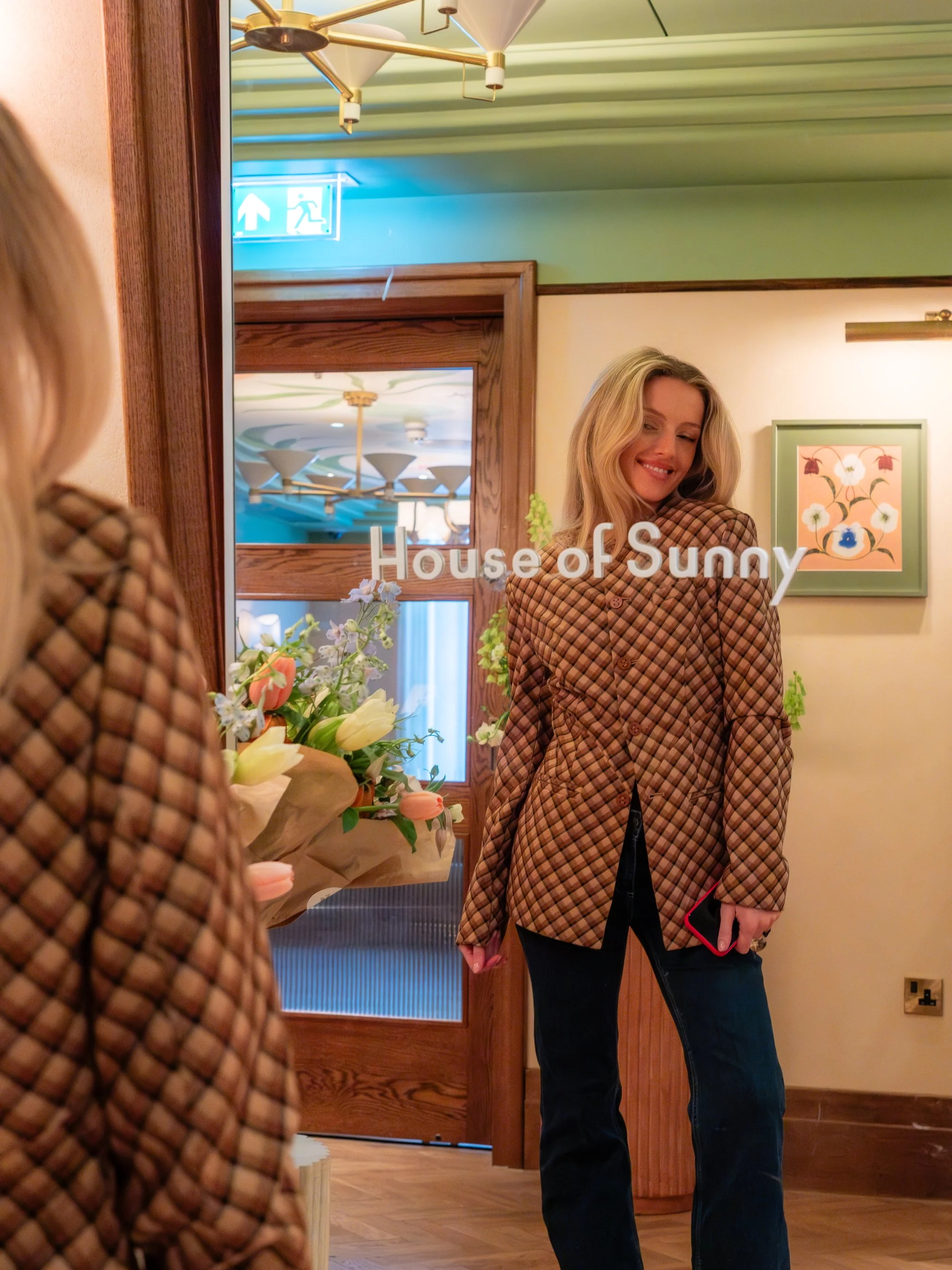 Reflection of a woman in a mirror with the words 'House of Sunny' on it, holding a red phone, standing in a warmly lit room decorated with flowers and framed art, with a green wall and wooden door frame in the background.