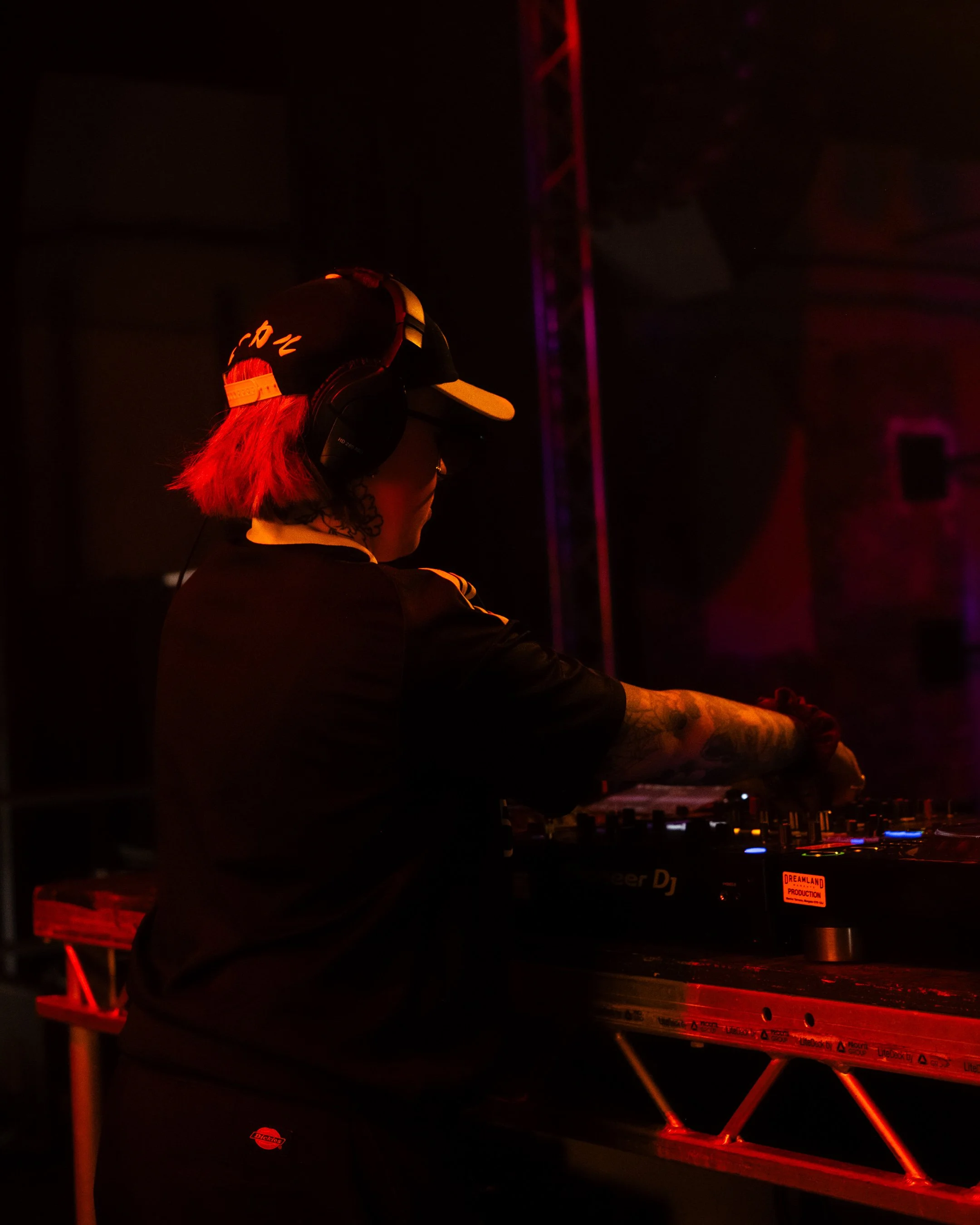 A DJ with red hair wearing headphones and a black cap labeled 'CR' performing at a dark nightclub or concert venue, illuminated by red and purple stage lights, with DJ equipment visible in front.