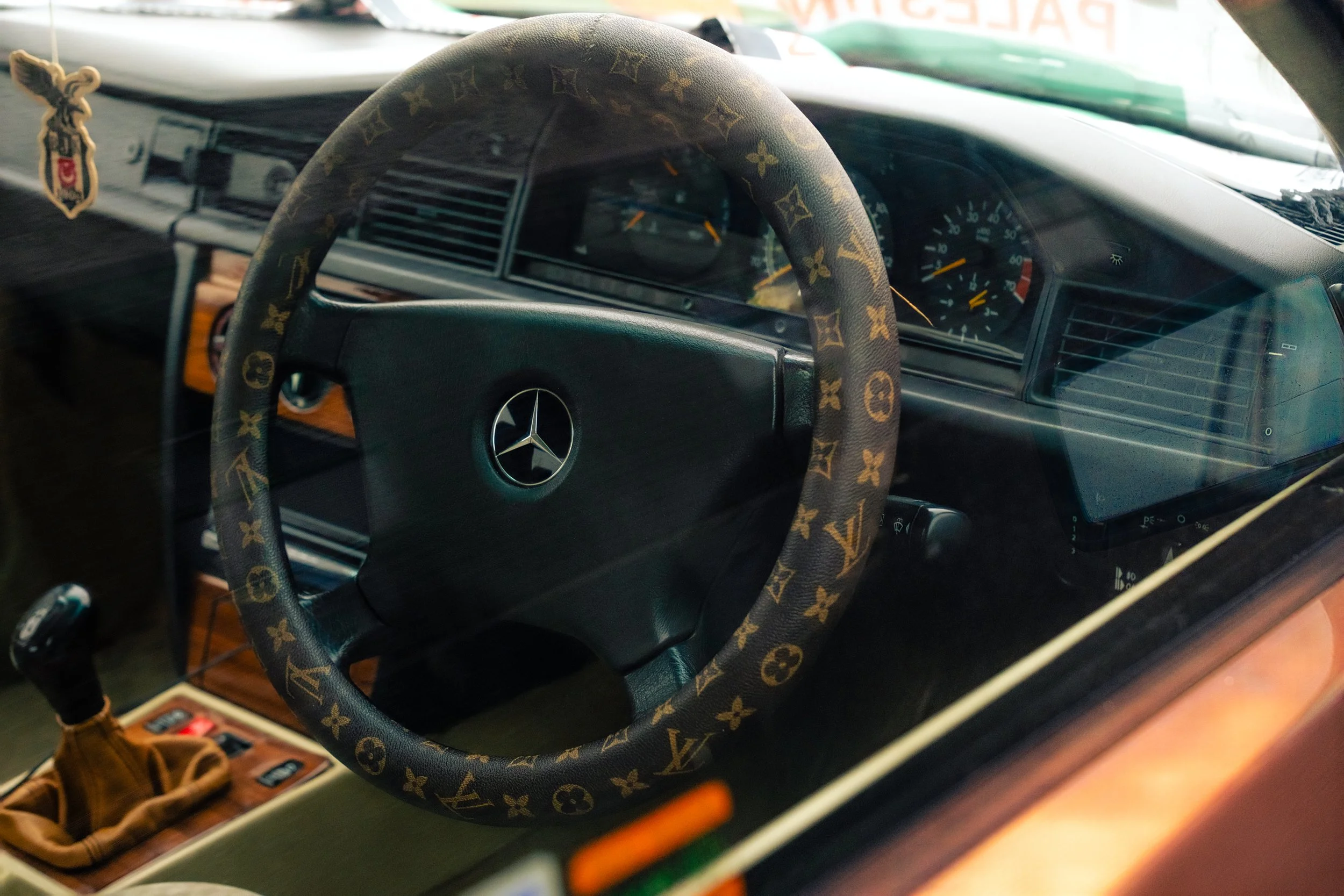 The interior of a car showing a steering wheel covered with a Louis Vuitton patterned cover, a dashboard with speedometer, and a dashboard console. Items like a bag and small objects are on the dashboard.
