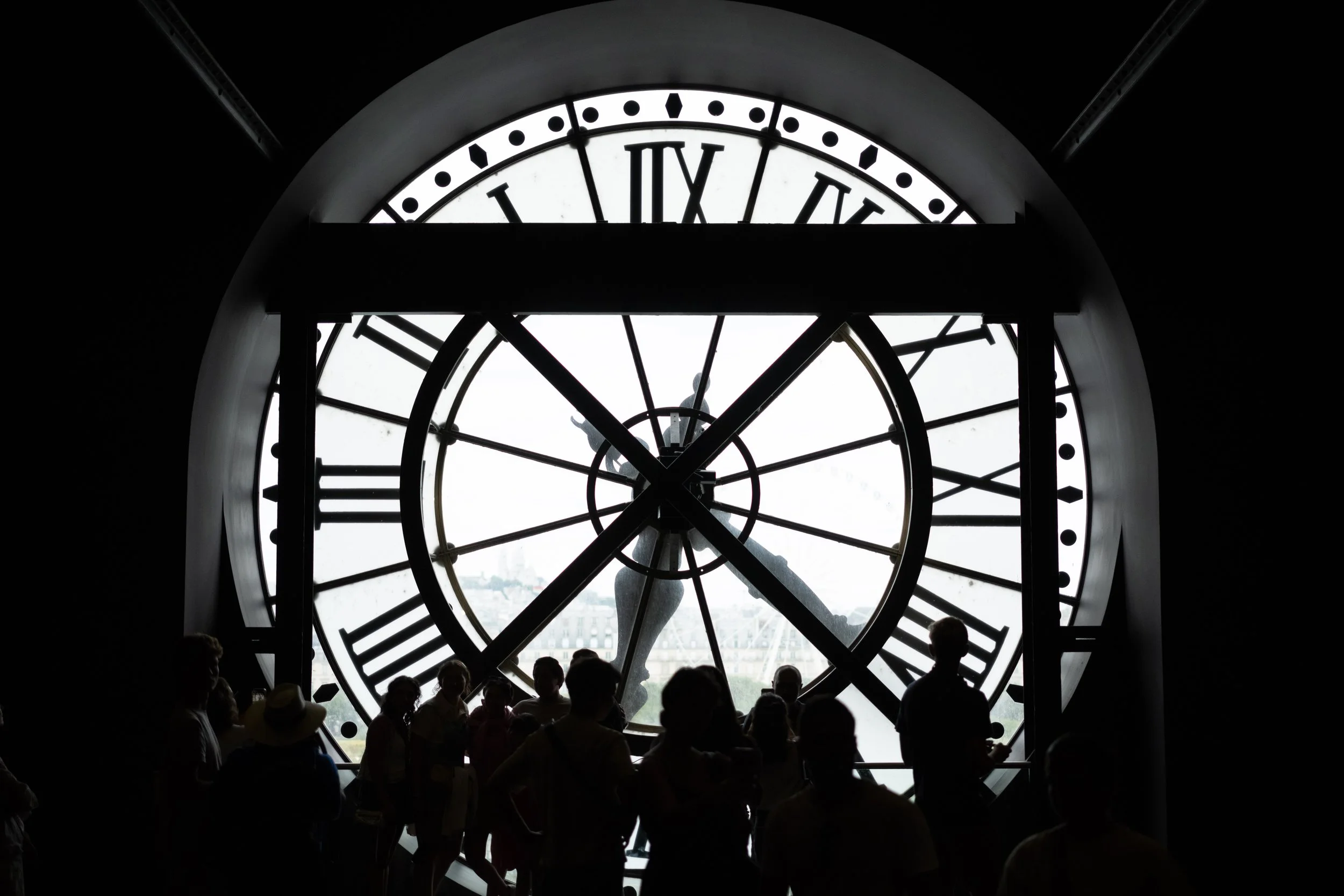 A large clock with Roman numerals viewed from inside a building, with silhouetted people standing in front of the clock face.