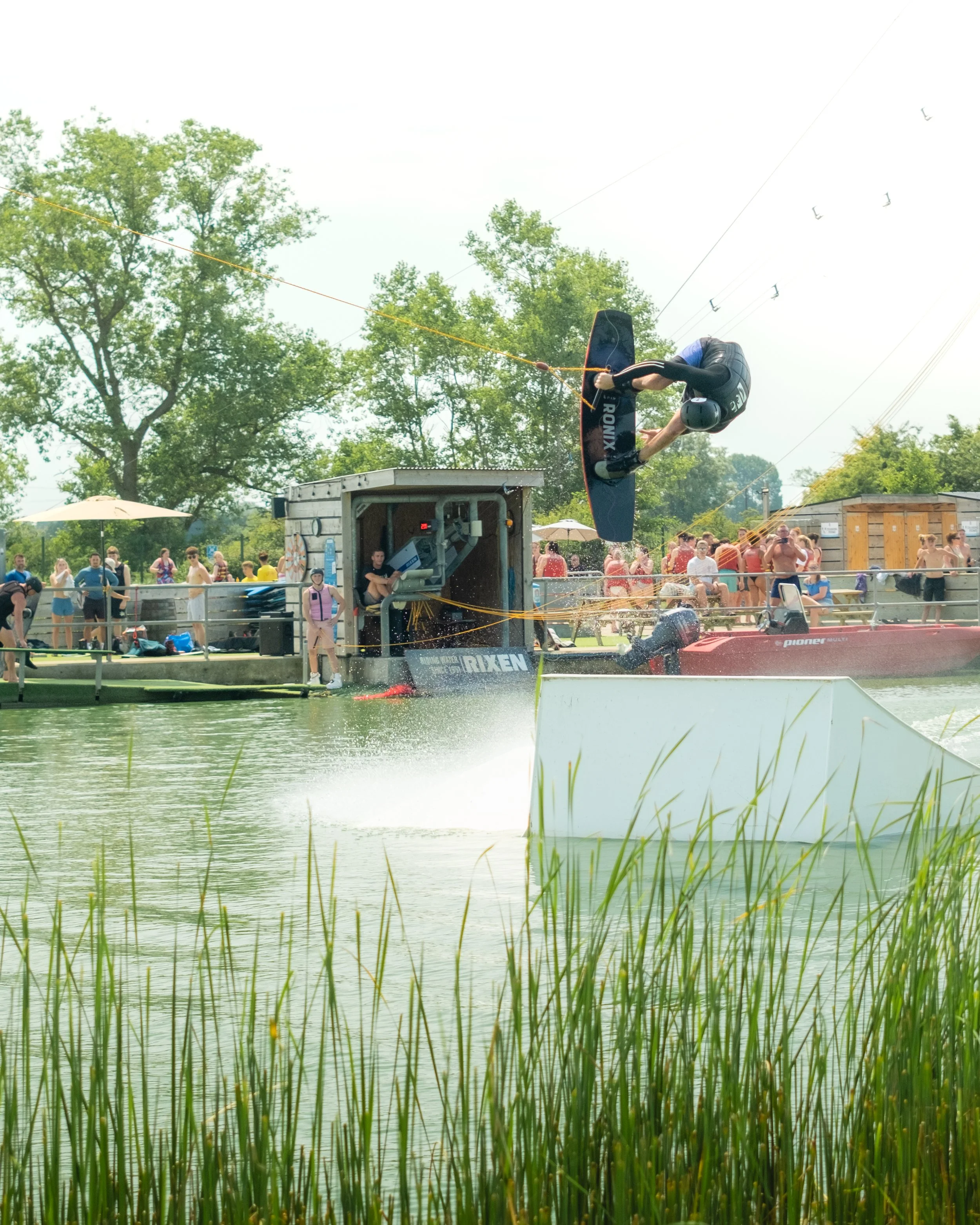 A person wakeboarding on a lake surrounded by trees during the daytime. The wakeboarder is mid-air after launching off a ramp, with water splashing below. Onlookers are watching on the shore.