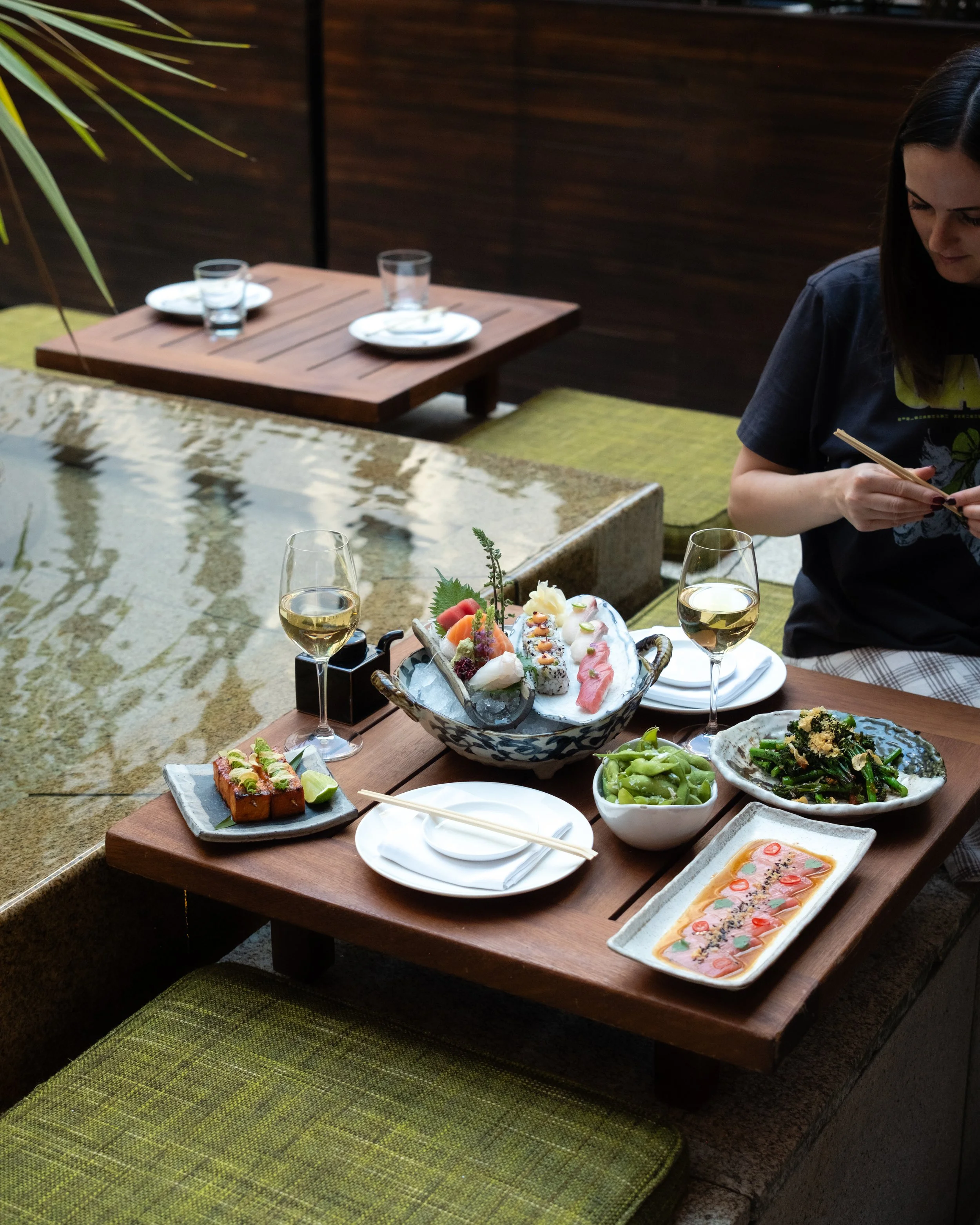 A table set with various Asian dishes, including sushi, edamame, garnished tofu, and a platter of colorful sashimi, with glasses of white wine, in an outdoor dining area with green cushions and wooden table.