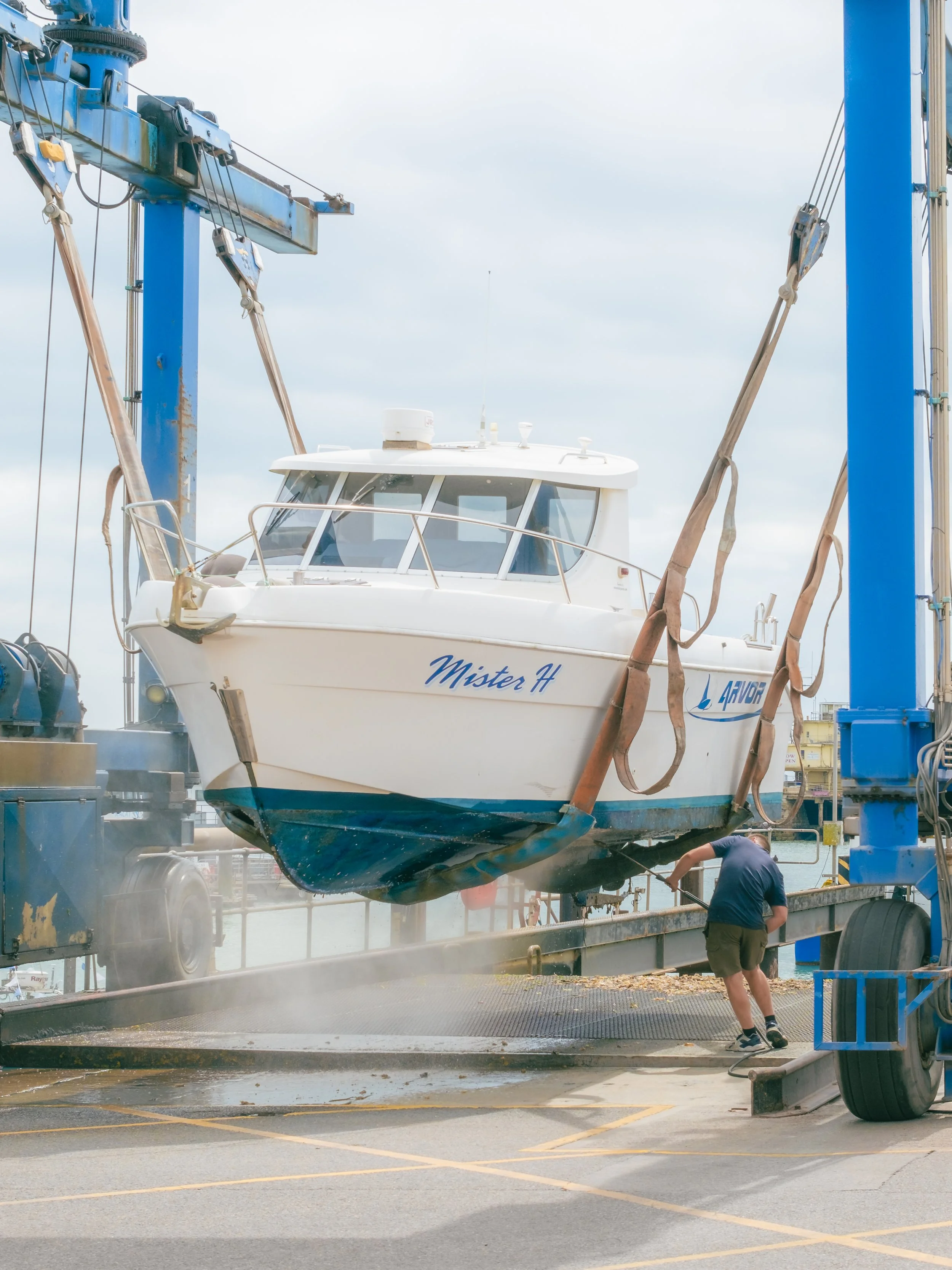 A man is working on a boat named Mister H that is suspended in the air by a lift at a boatyard. The boat is being washed or cleaned, creating a mist around it.