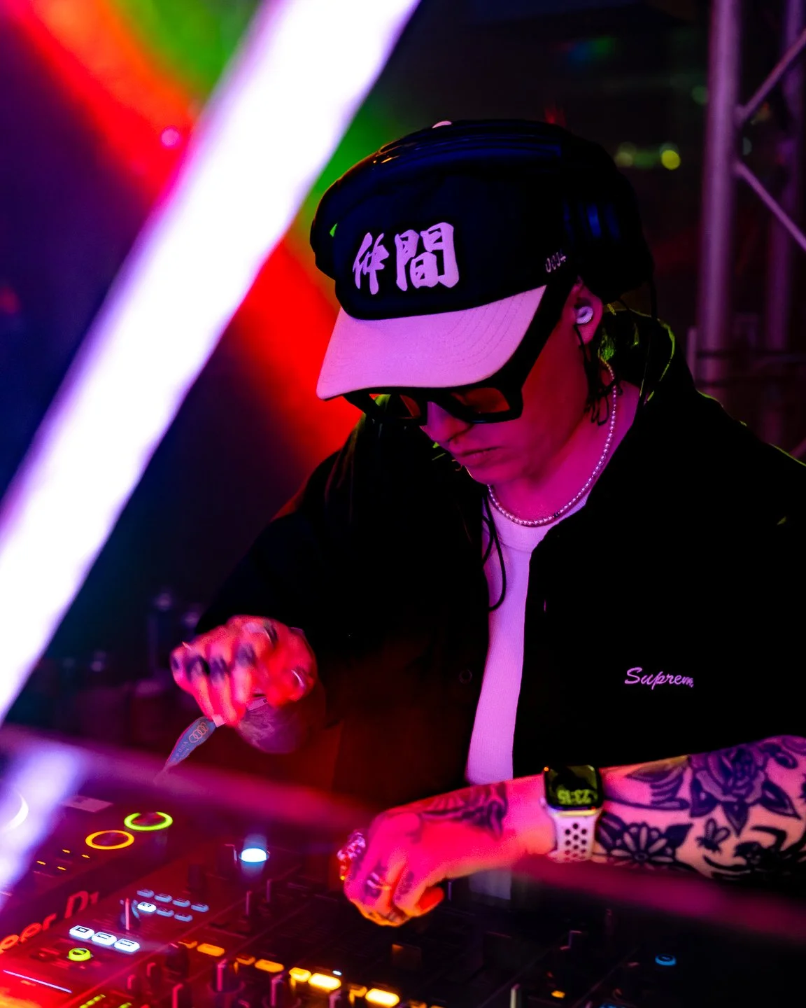 A DJ wearing a cap with Japanese characters, sunglasses, and jewelry, operating DJ equipment in a colorful, neon-lit environment.