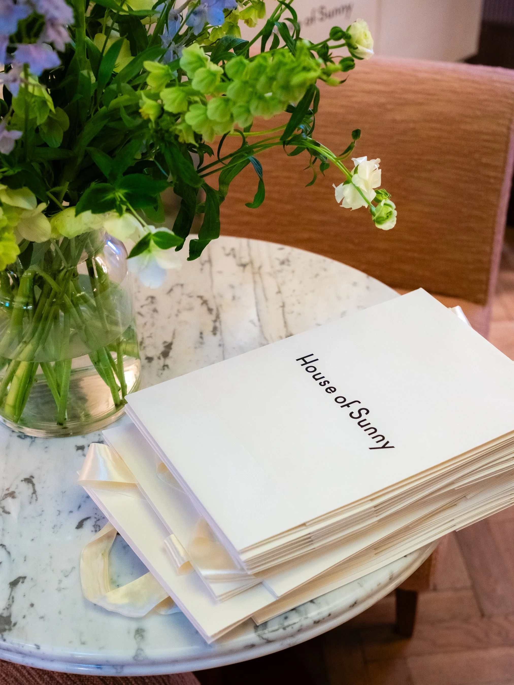 Close-up of a stack of papers on a table with a bouquet of green and purple flowers in a glass vase nearby.