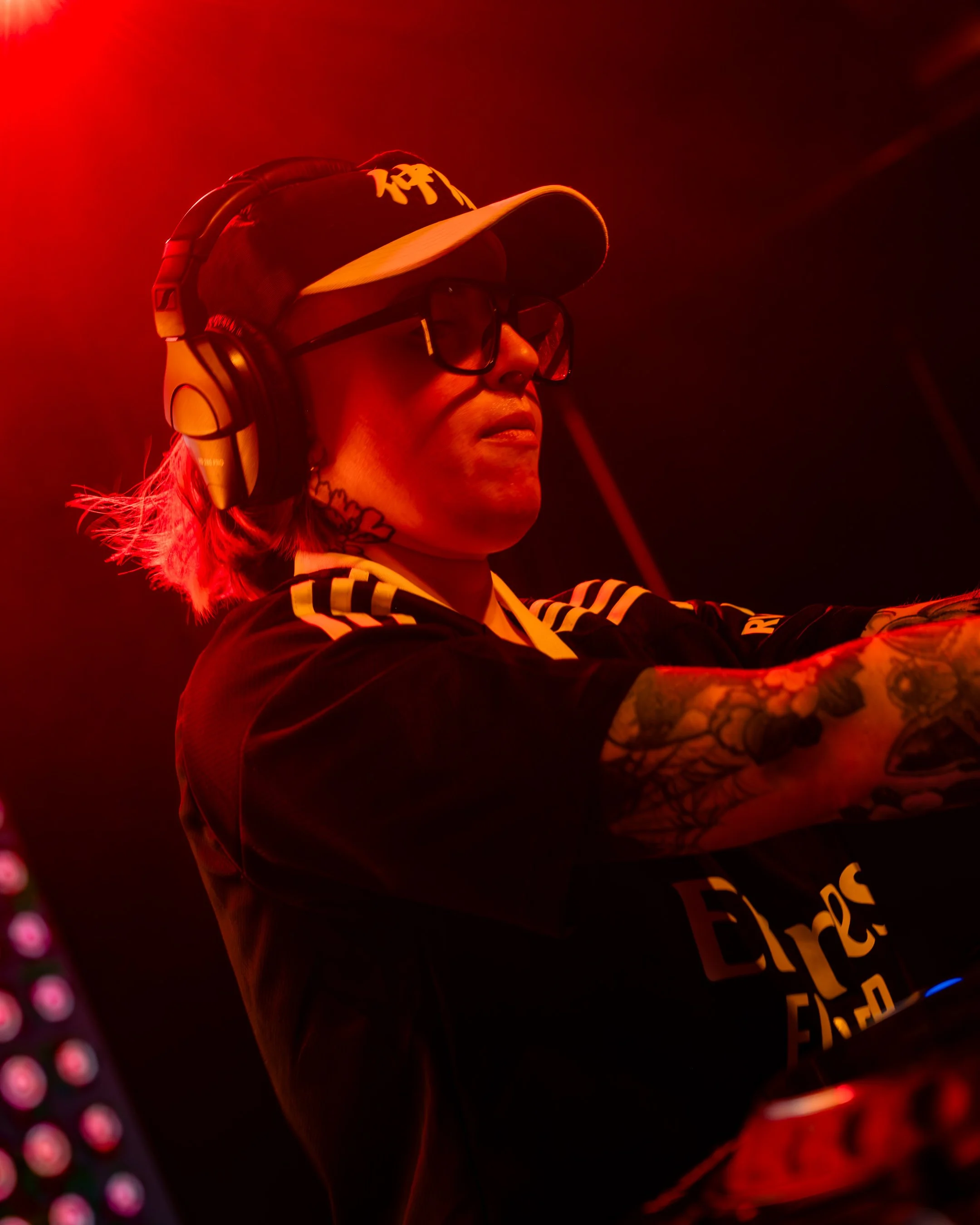 A woman wearing a dark baseball cap, sunglasses, headphones, and a black shirt with yellow text, standing in a red-lit environment.