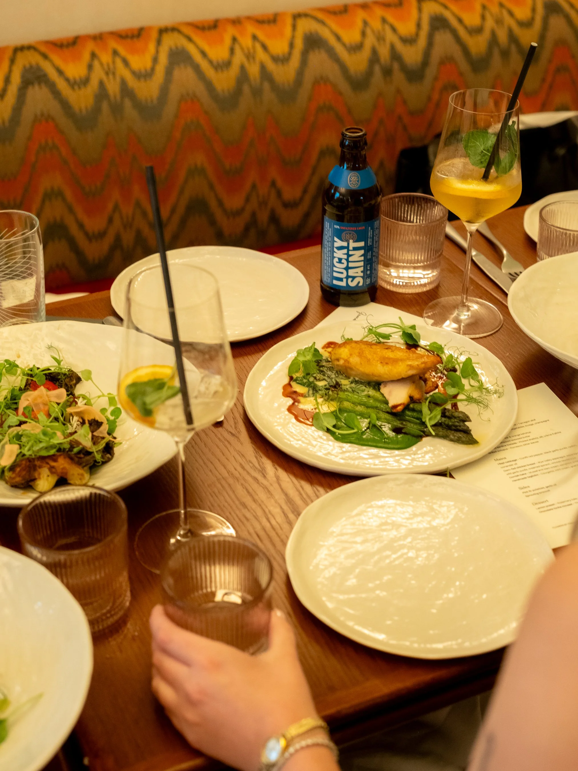 A dining table set with plates and glasses, with a hand holding a glass of dark beverage in the foreground. There is a bottle of Lucky Saint non-alcoholic beer, a cocktail with mint and a straw, and a plate of grilled chicken with vegetables.