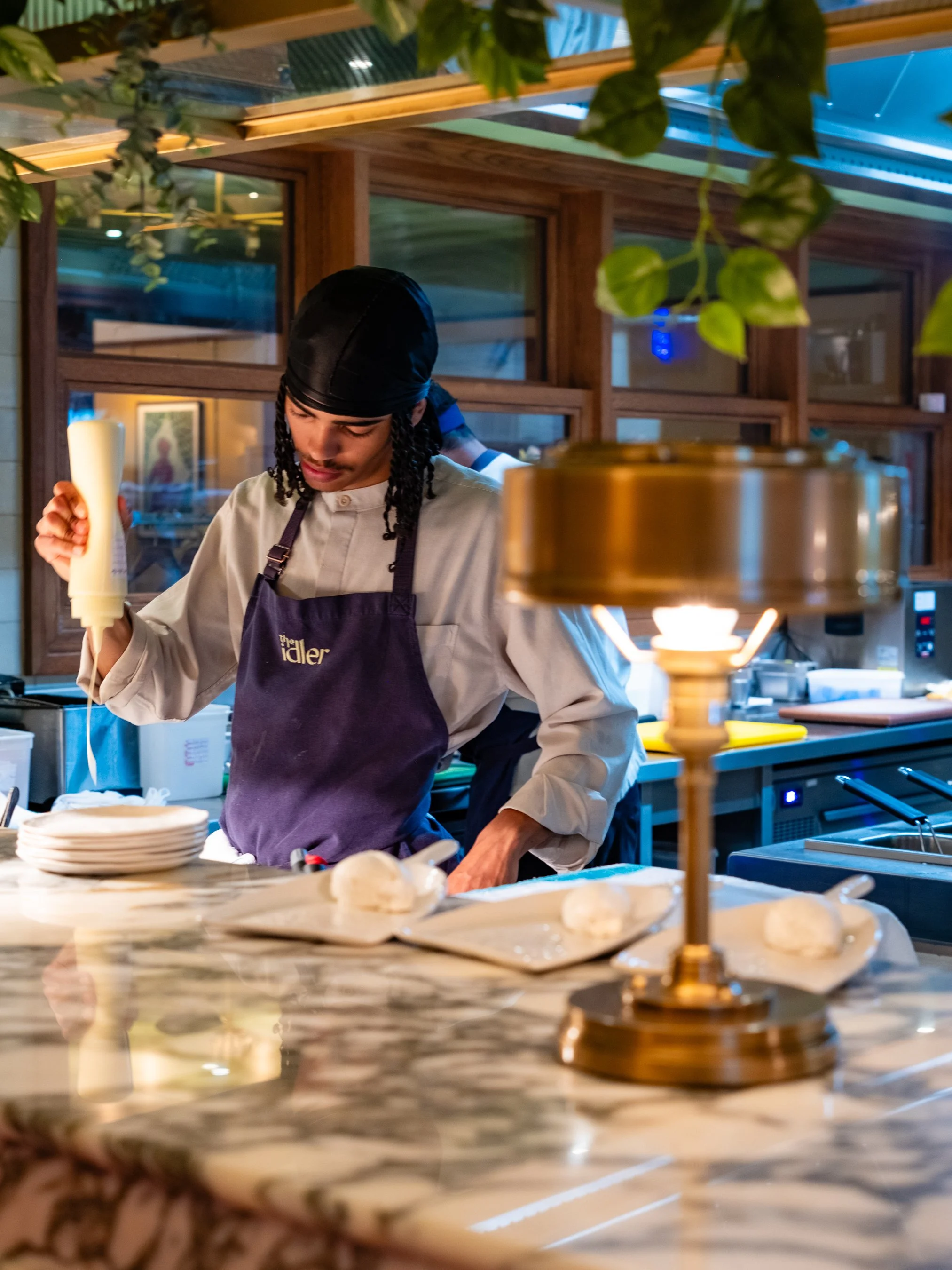 A chef in a black hair covering and purple apron preparing food in a restaurant kitchen with a marble countertop, plates, and a golden candle holder in the foreground.