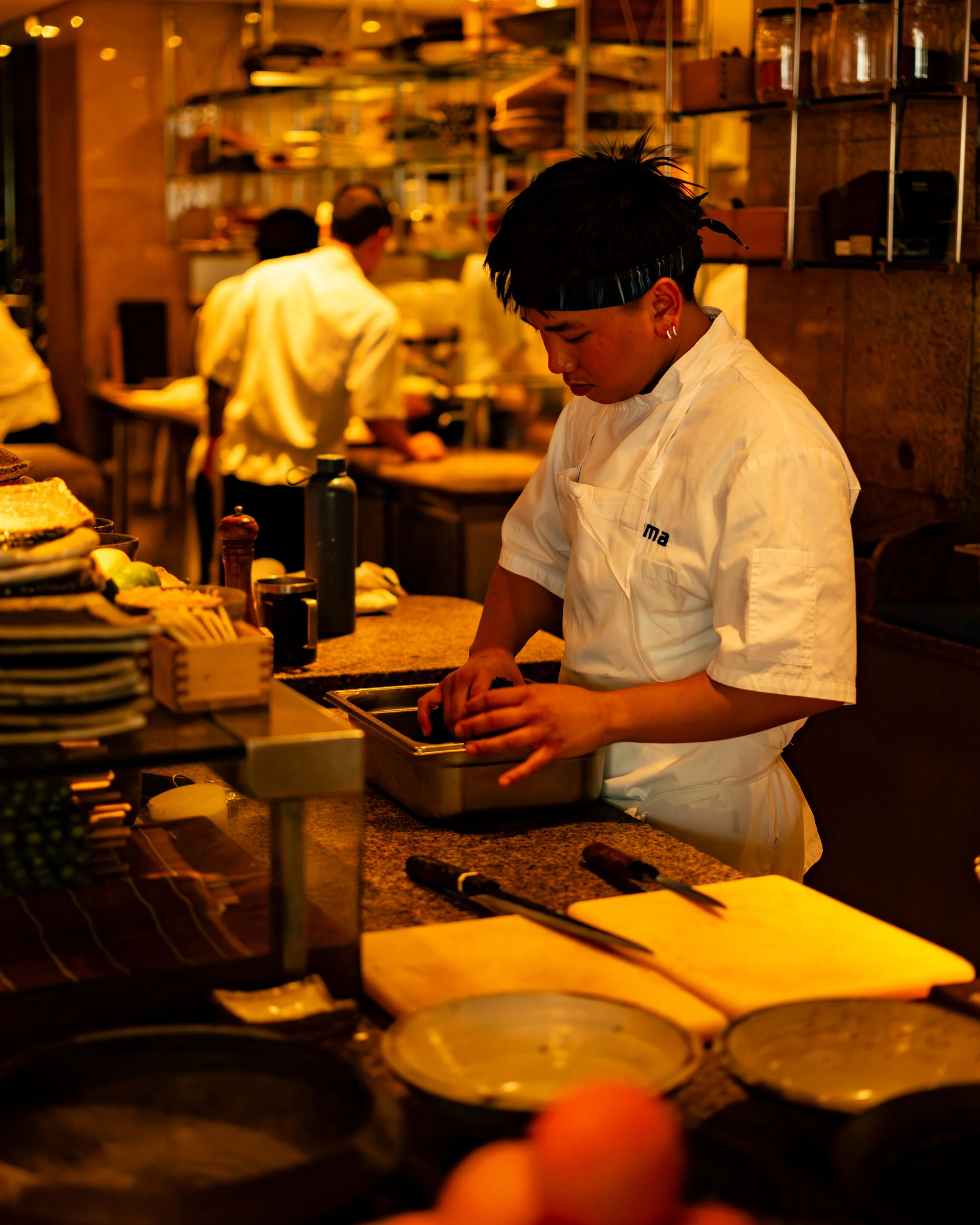 A chef with short dark hair and earrings working in a warm-lit restaurant kitchen. Two more chefs are in the background. The counter is cluttered with plates, utensils, and ingredients under a shelf with jars and kitchen tools.