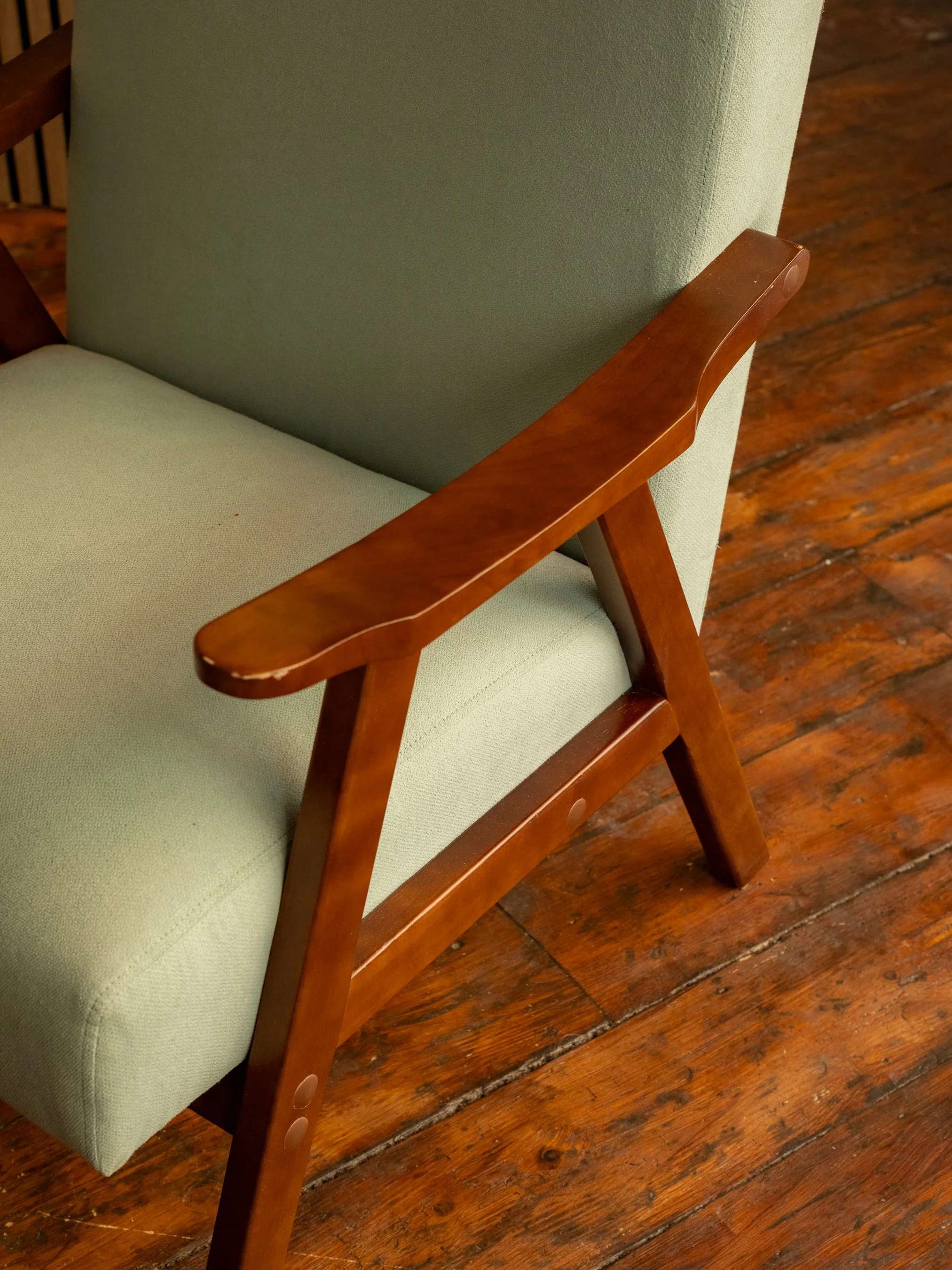 Close-up of a wooden arm of a chair with light green fabric upholstery and a matching cushion, on a wooden floor.