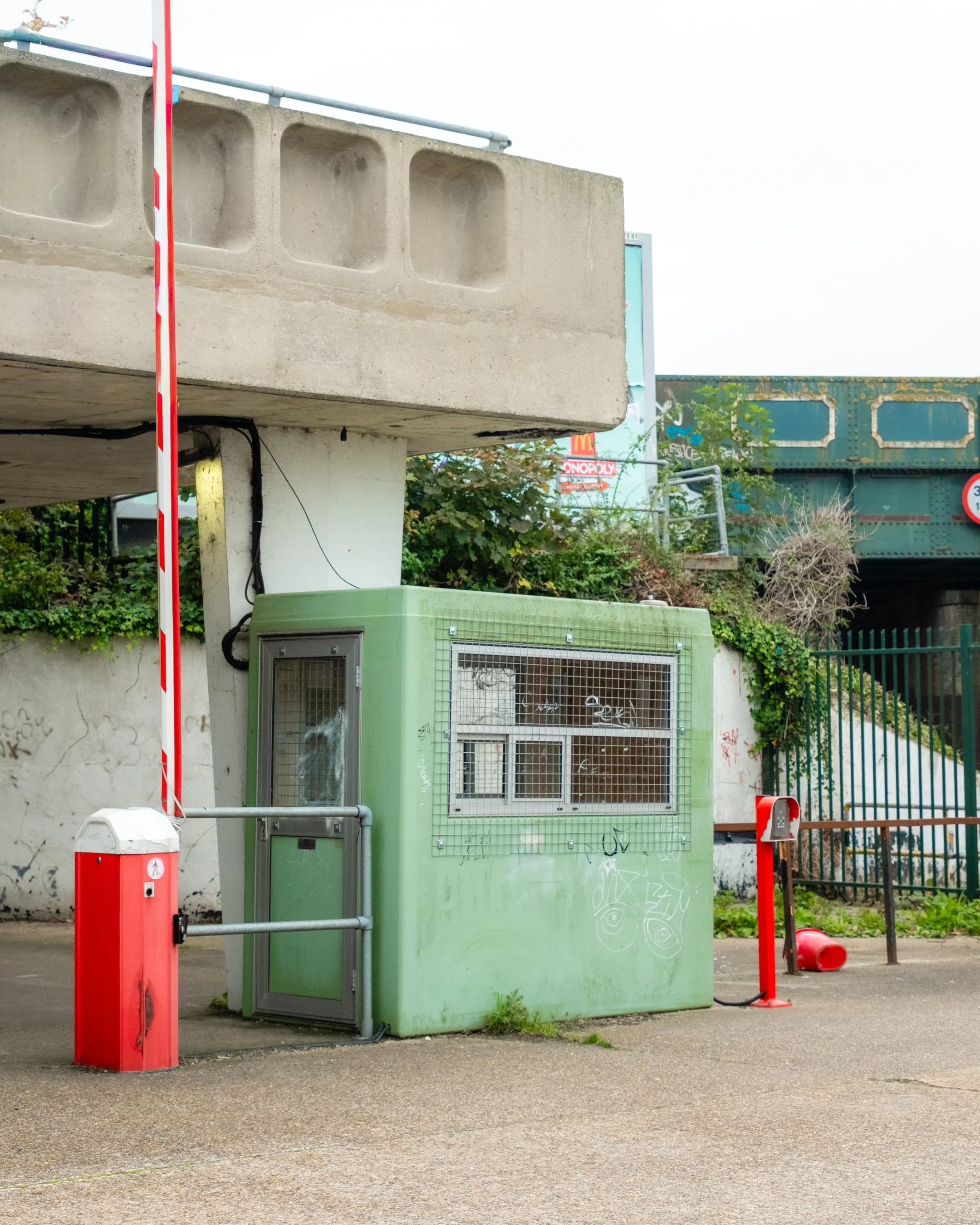 A small guard booth painted green is located at a parking lot entrance, with graffiti on its surface. Next to it is a red and white barrier gate, and there is a red trash bin tipped over on the ground. In the background, there is a large green train 