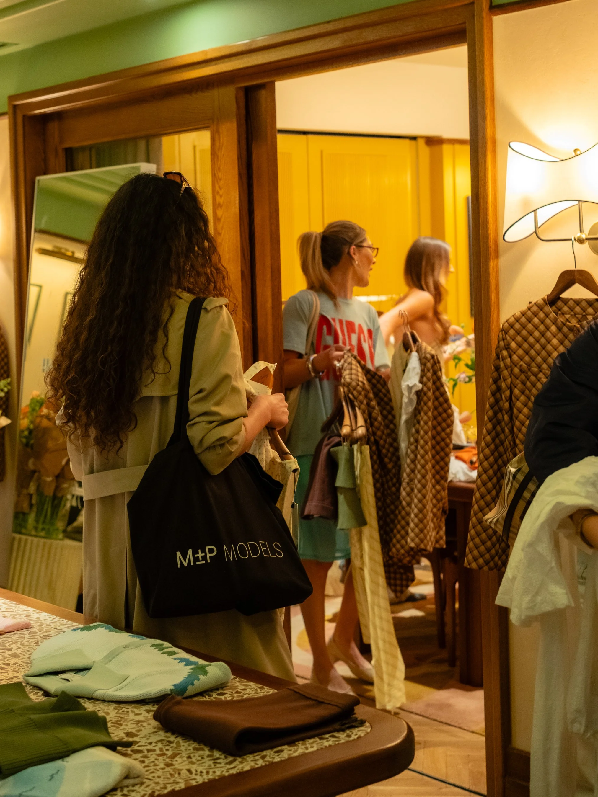 People shopping for folded clothes in a store, with a table of clothing items in the foreground and a mirror reflecting some individuals in the background.