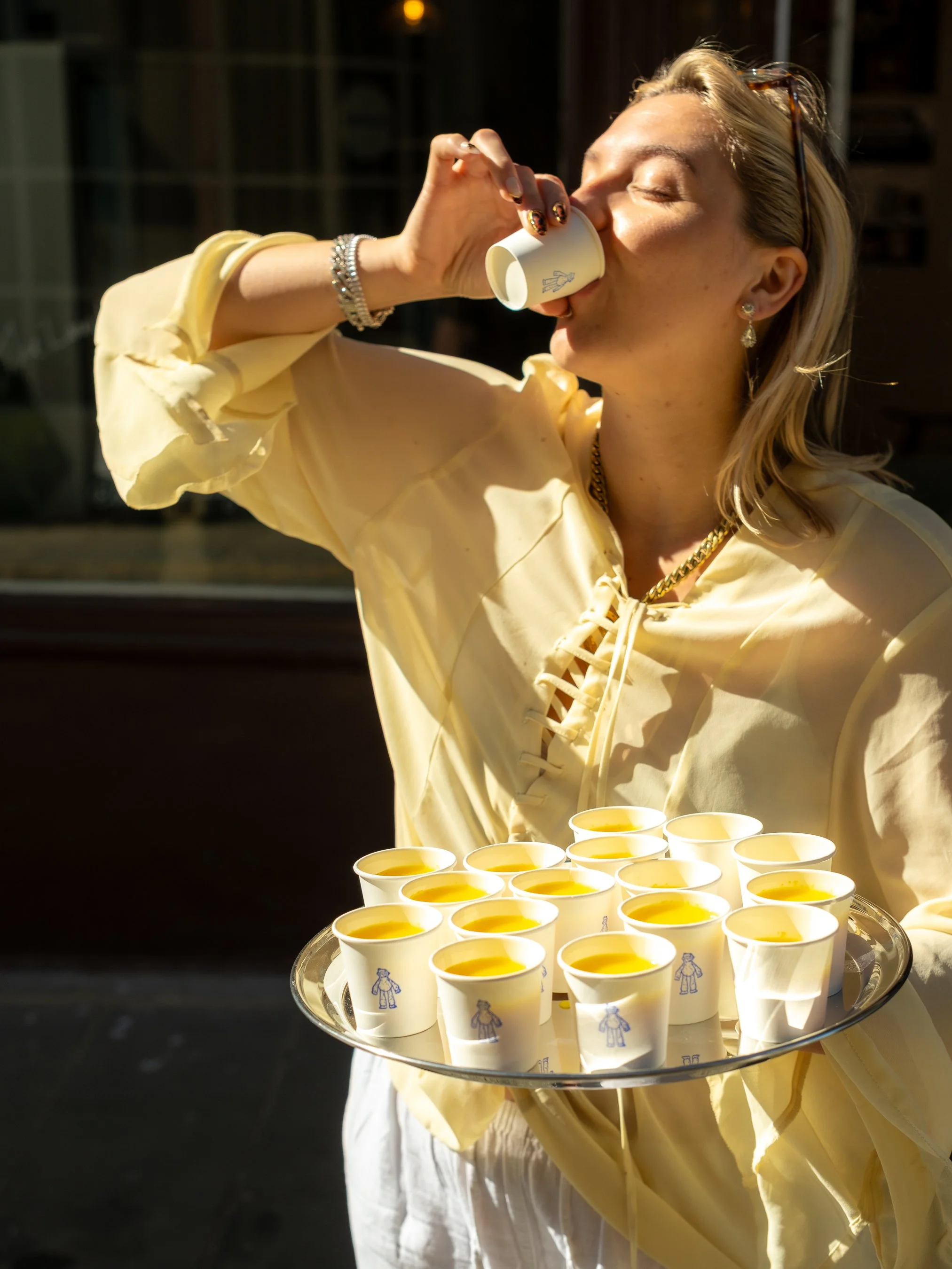 A woman in a yellow dress with jewelry is holding a tray with small cups of yellow liquid, possibly juice, and sipping from a cup.