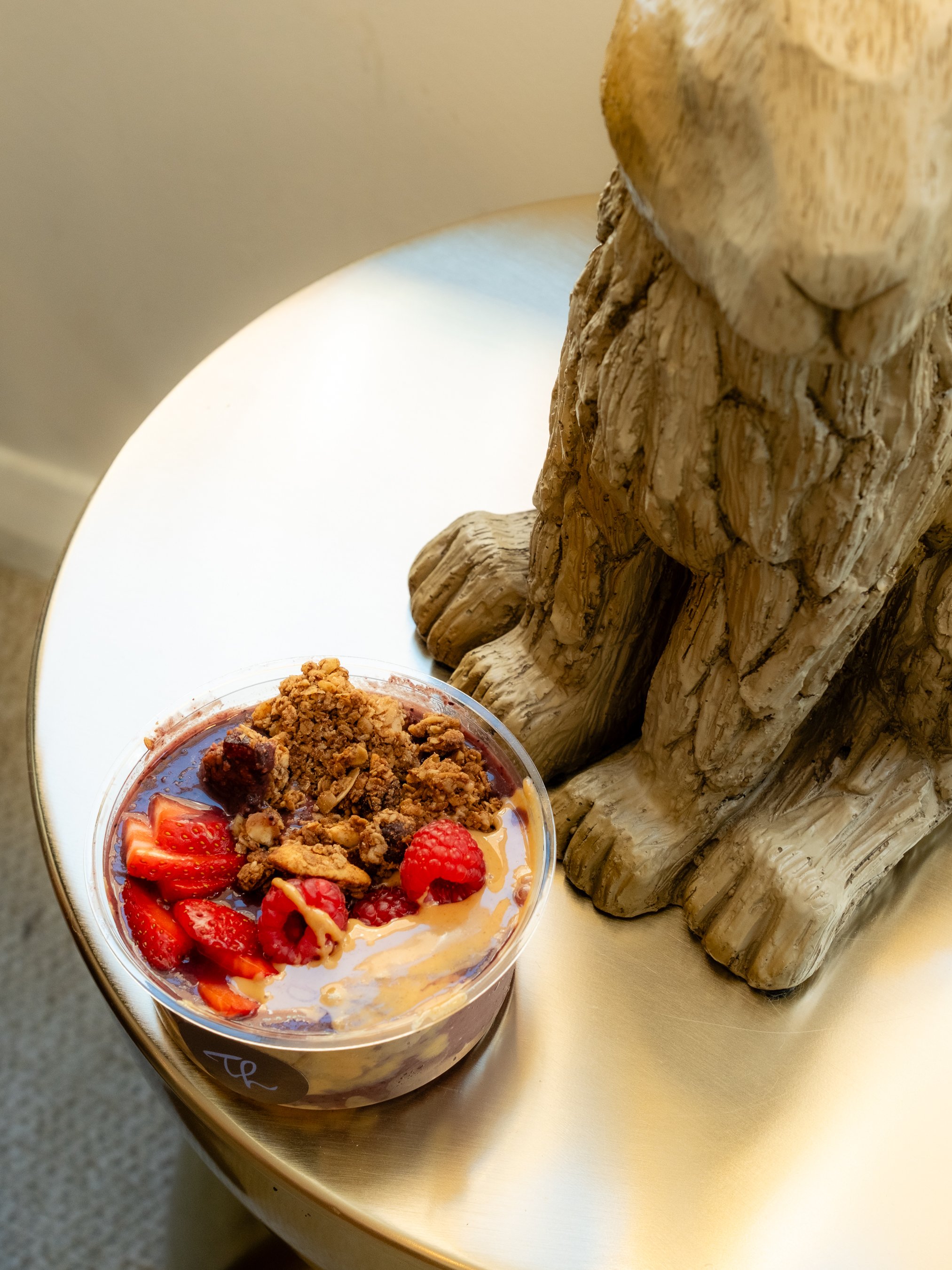A dessert bowl filled with strawberries, raspberries, granola, and syrup, placed on a gold-colored table next to a decorative wooden lion sculpture.
