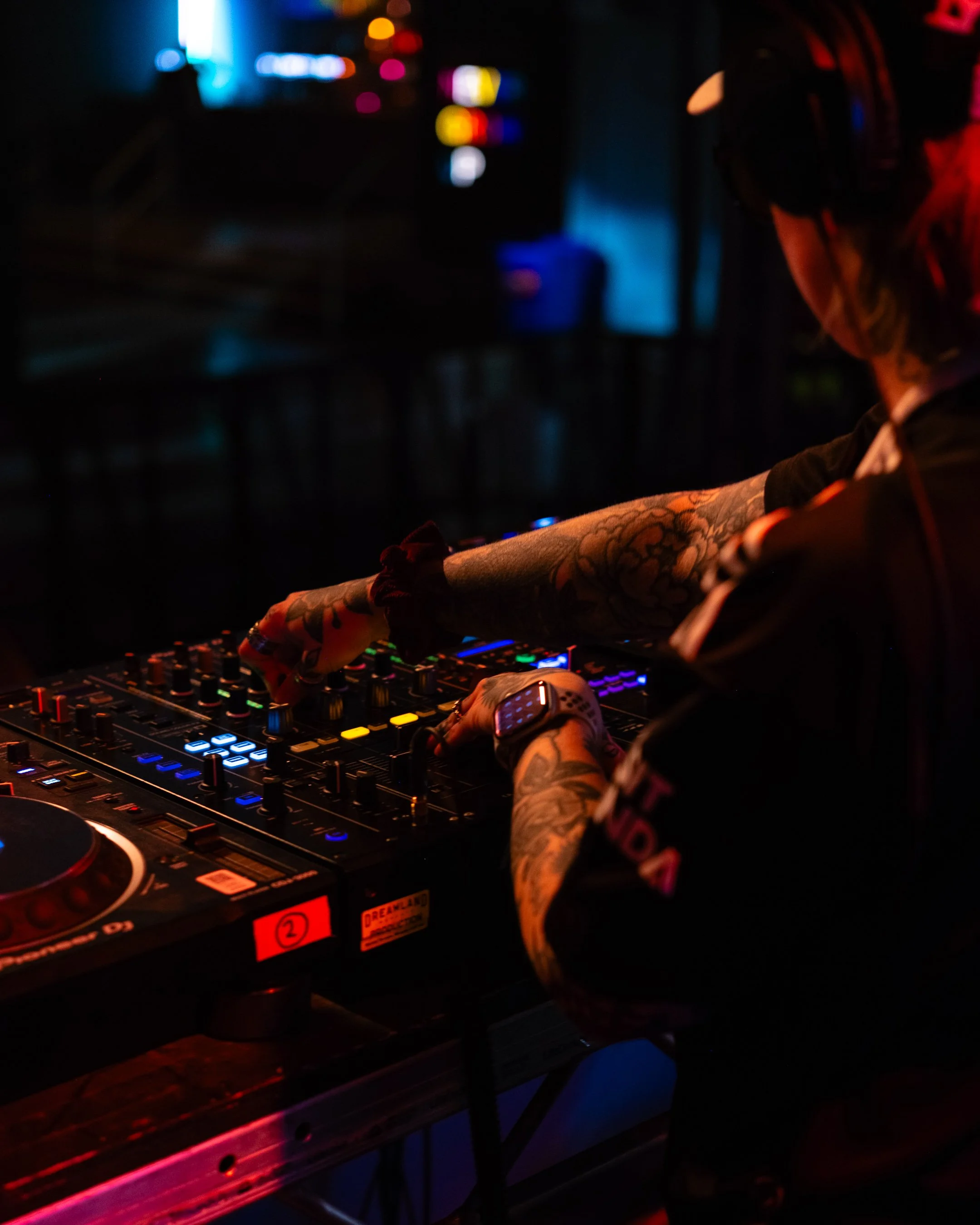 A DJ with colorful tattoos on arms is mixing music on a DJ console at a nightclub, with a dark background and blue and red lighting.