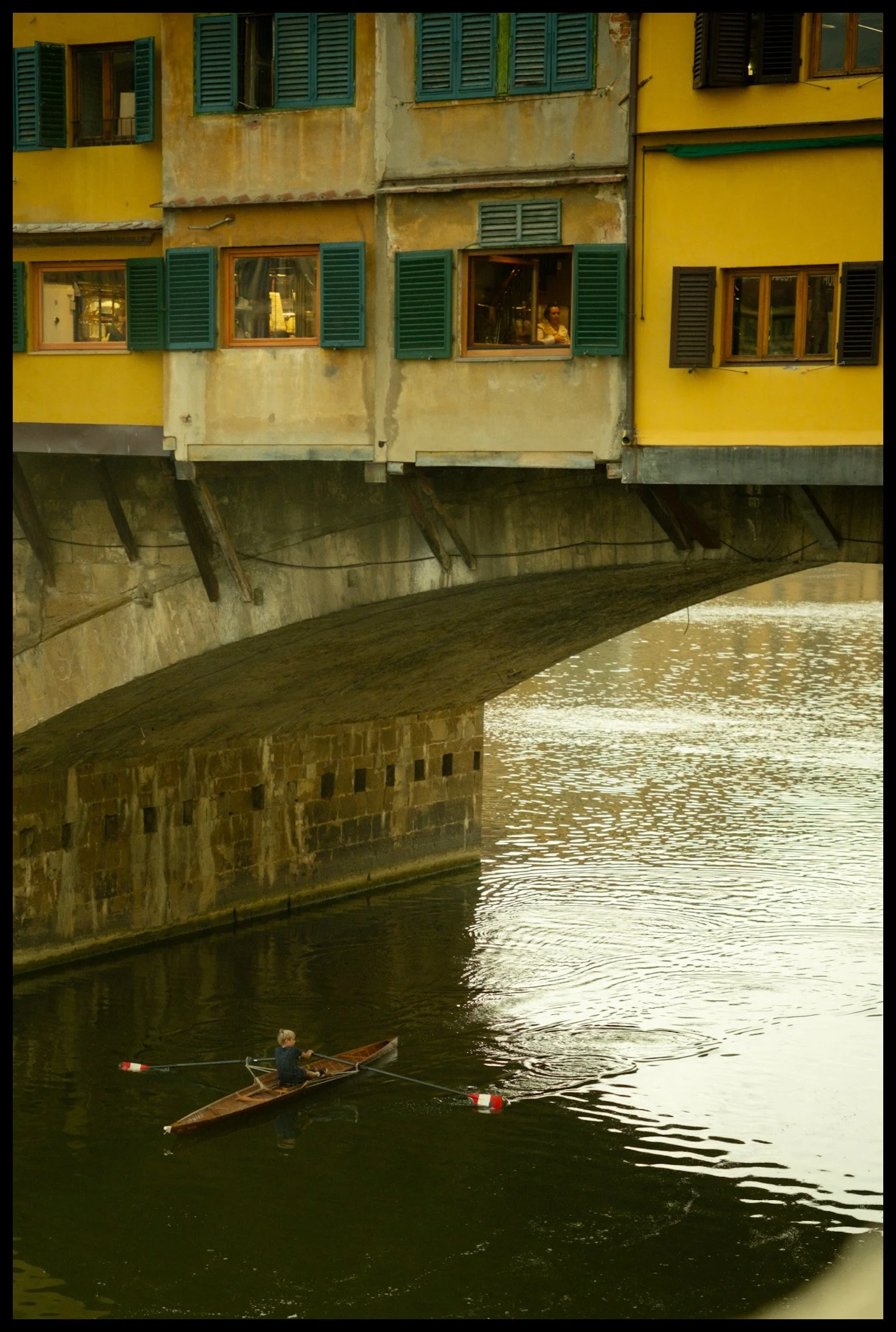 A person row boating under a bridge with colorful, weathered buildings with open windows, some with shutters, along a river. The building has a yellow facade and green shutters.