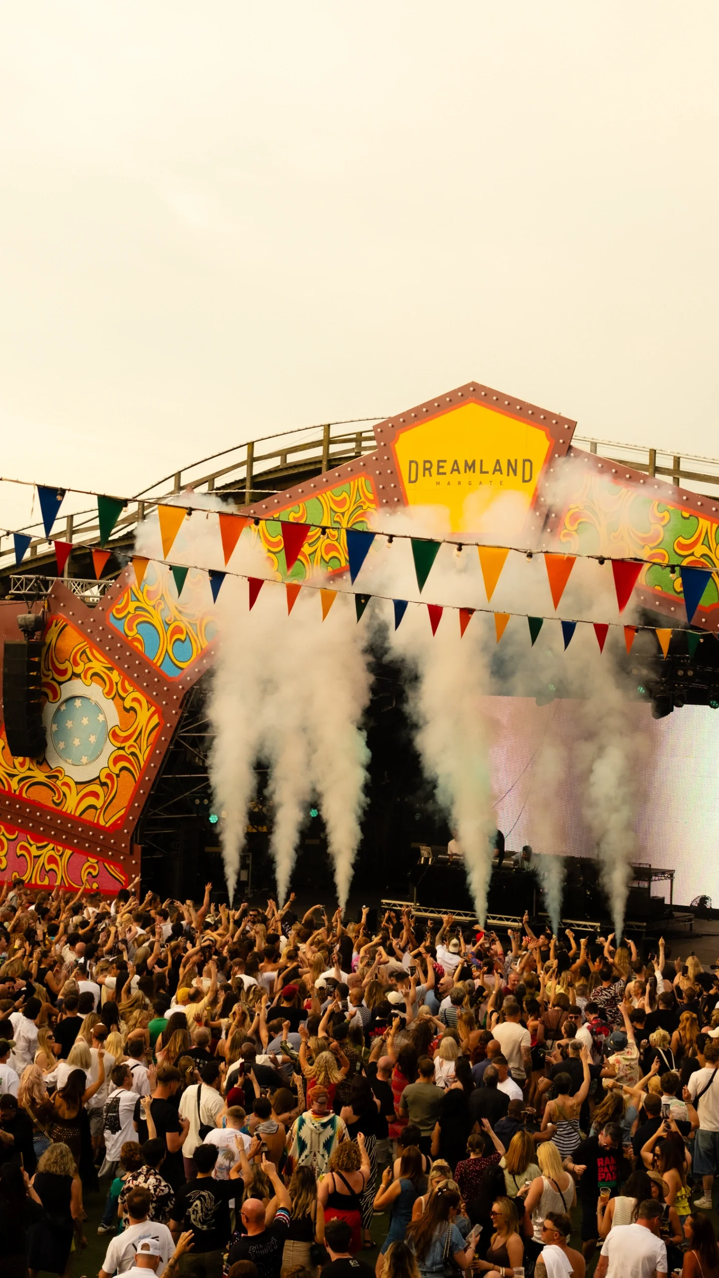 Crowd at an outdoor concert with stage, colorful decorations, fog, and smoke effects, and a sign reading 'Dreamland Margate'.