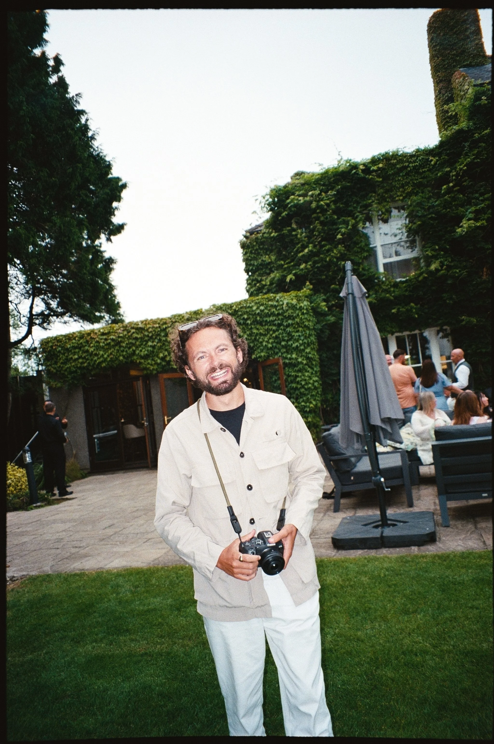 A smiling man with dark curly hair and a beard holding a camera stands outdoors in front of a green lawn. In the background, there are people gathered near a patio area with lounge chairs, a large umbrella, and lush building covered in green ivy.