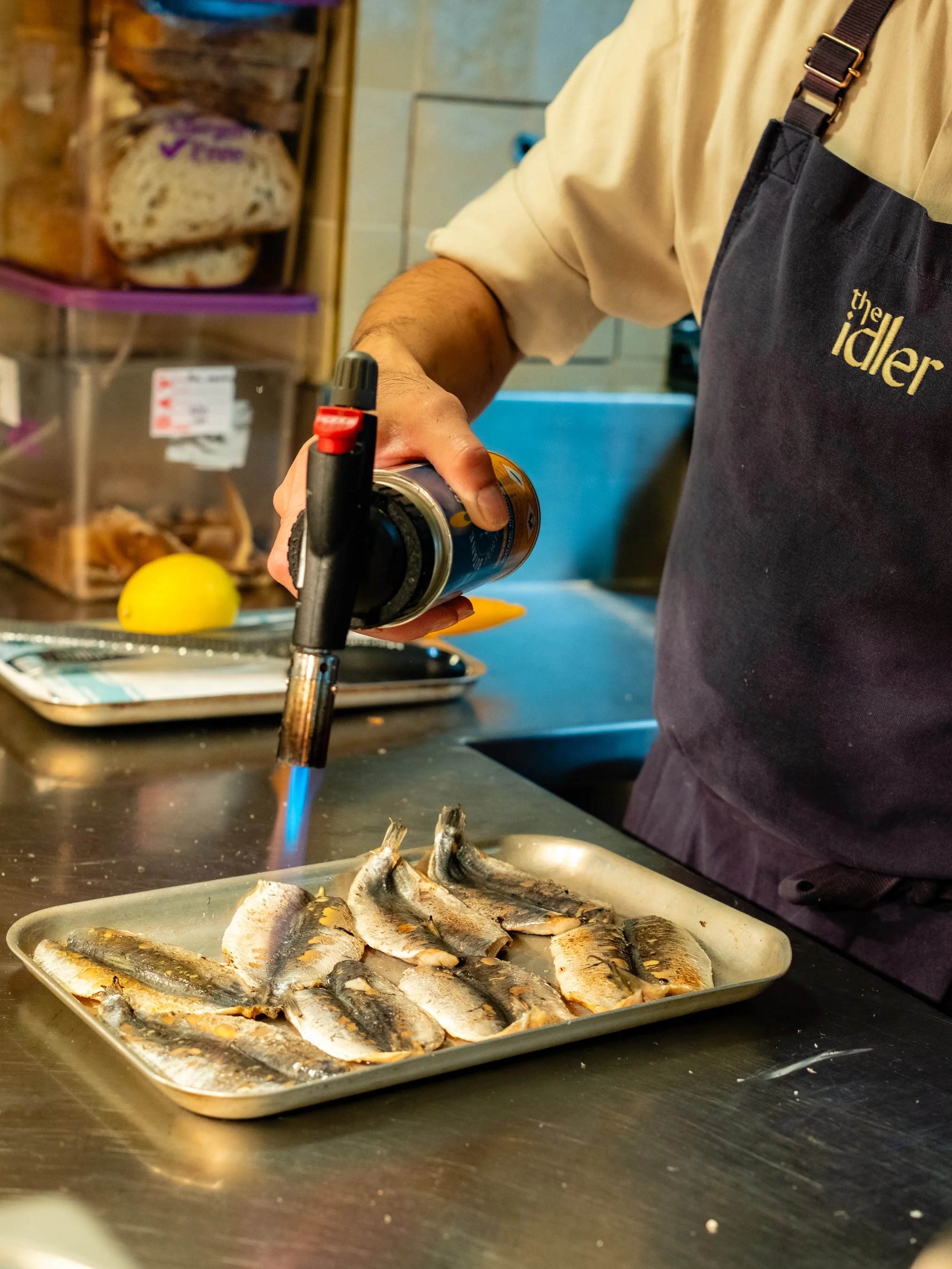 A person wearing a black apron with the word 'the idler' on it is holding a can of aerosol and using a torch to cook small fish on a tray, with a tray of cooked fish and yellow lemon nearby.