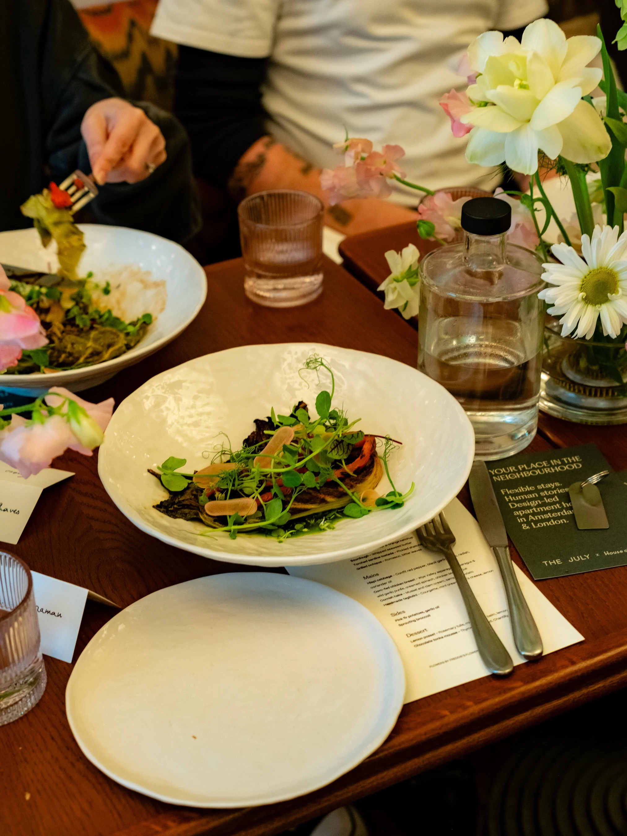 Table setting with white plates, a glass of water, a bottle of water, a fork and a knife, a menu, and a pink and white flower arrangement. Food on some plates, including a dish with greens and a cooked item with herbs.