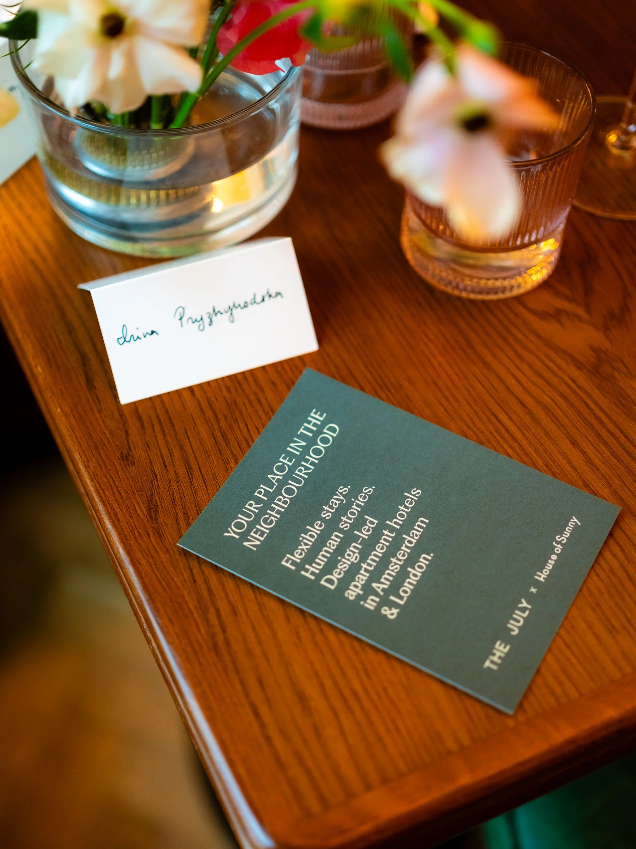 A wooden table with a flower arrangement in a clear glass vase and pink glasses. A card with handwritten text is visible, with printed material partially seen in the corner.