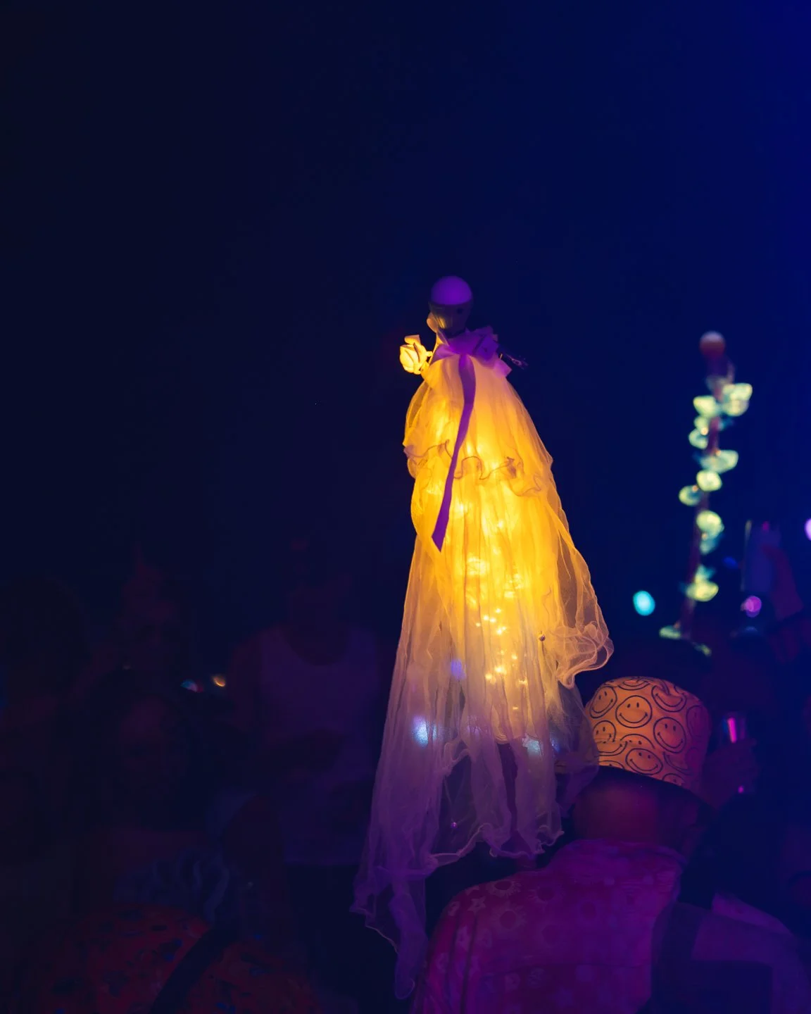 A mannequin dressed in a glowing, yellowish-orange gown with lights inside, illuminated under dark lighting, at a display or event.