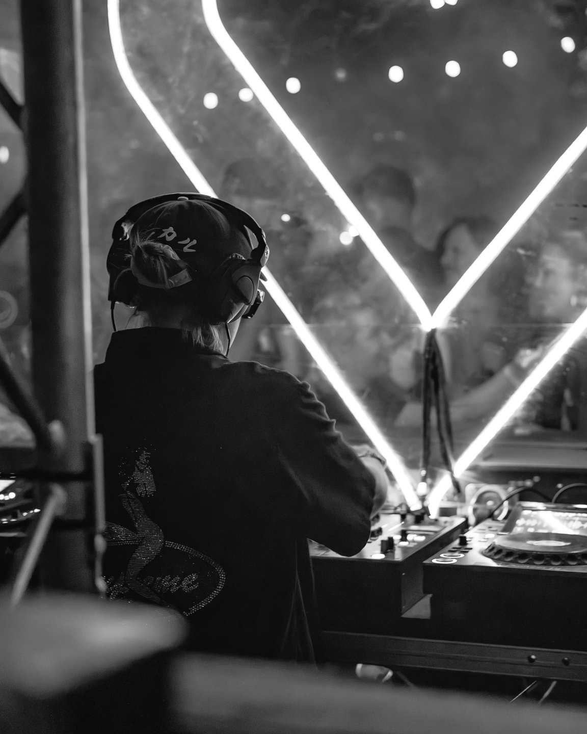A DJ wearing headphones and a cap, standing behind a DJ booth with musical equipment, illuminated by geometric light beams in a dark setting.