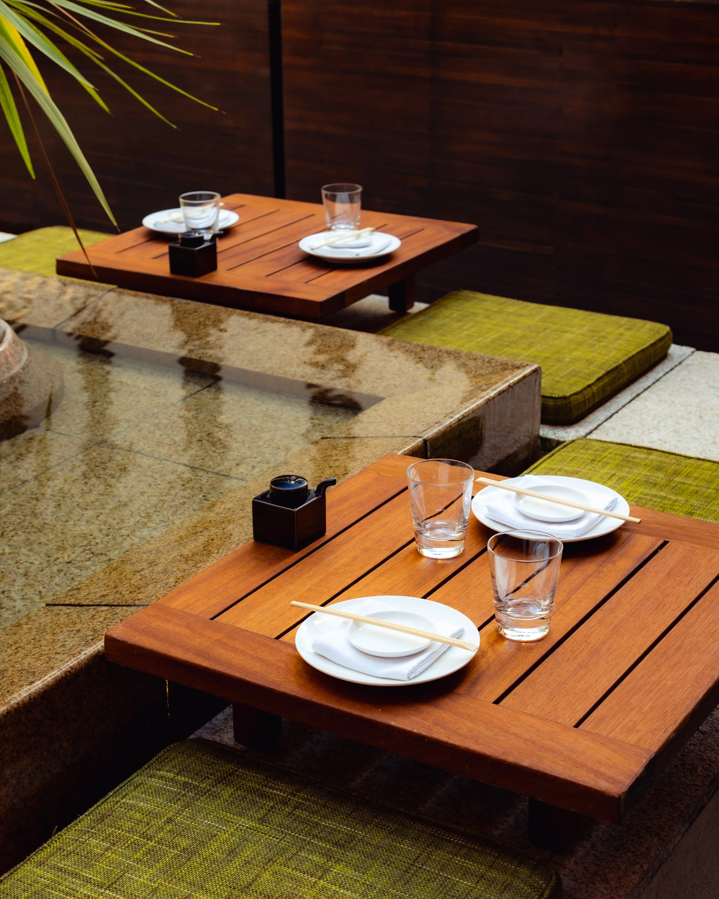 A wooden table set for dining with plates, glasses, chopsticks, and a small black container, surrounded by green cushions on the floor, in an outdoor or patio area with a dark wooden wall and a plant with long leaves nearby.