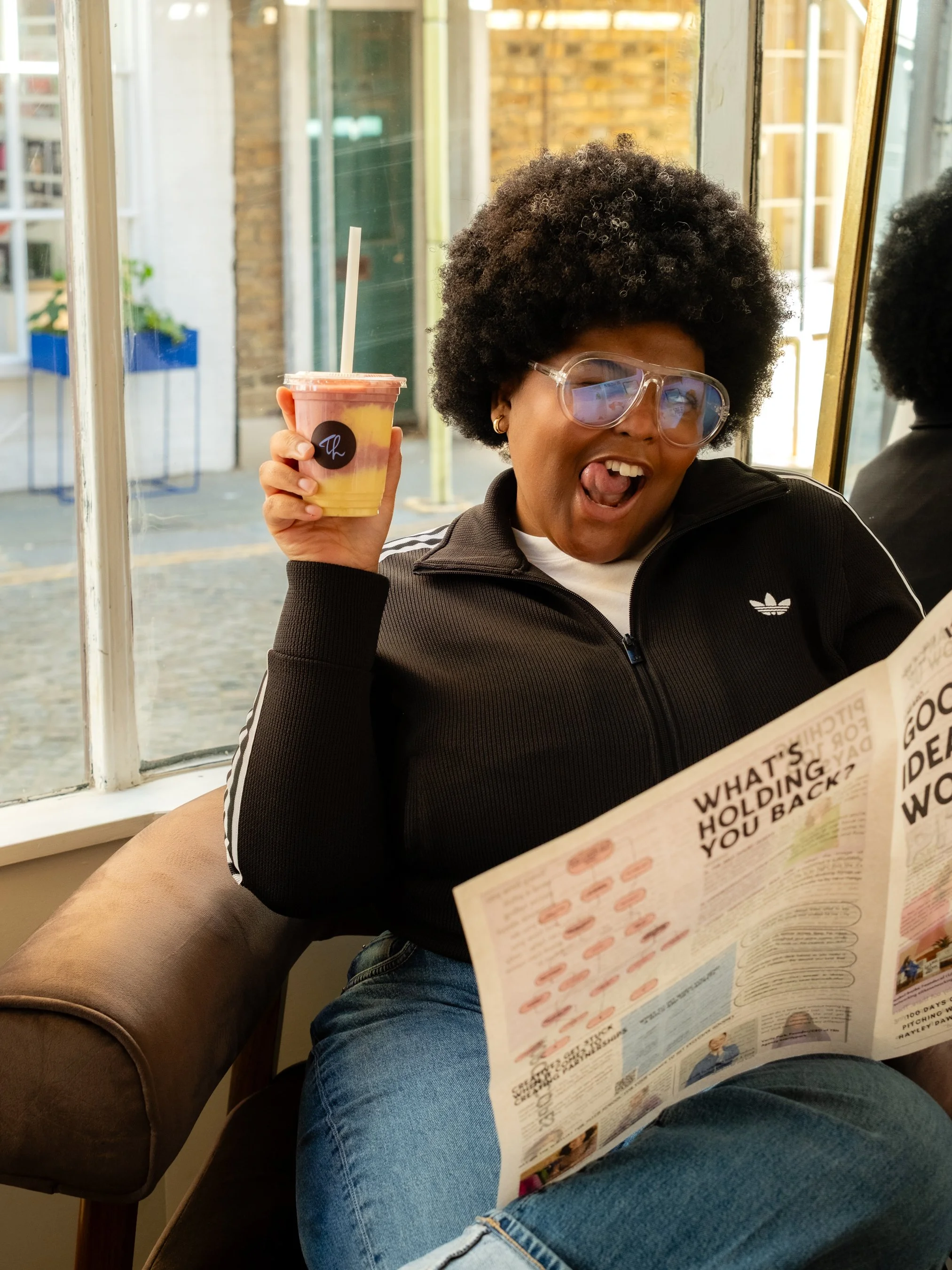 A woman with curly hair and glasses holding a smoothie and sticking out her tongue, sitting at a café table with a menu in front of her.
