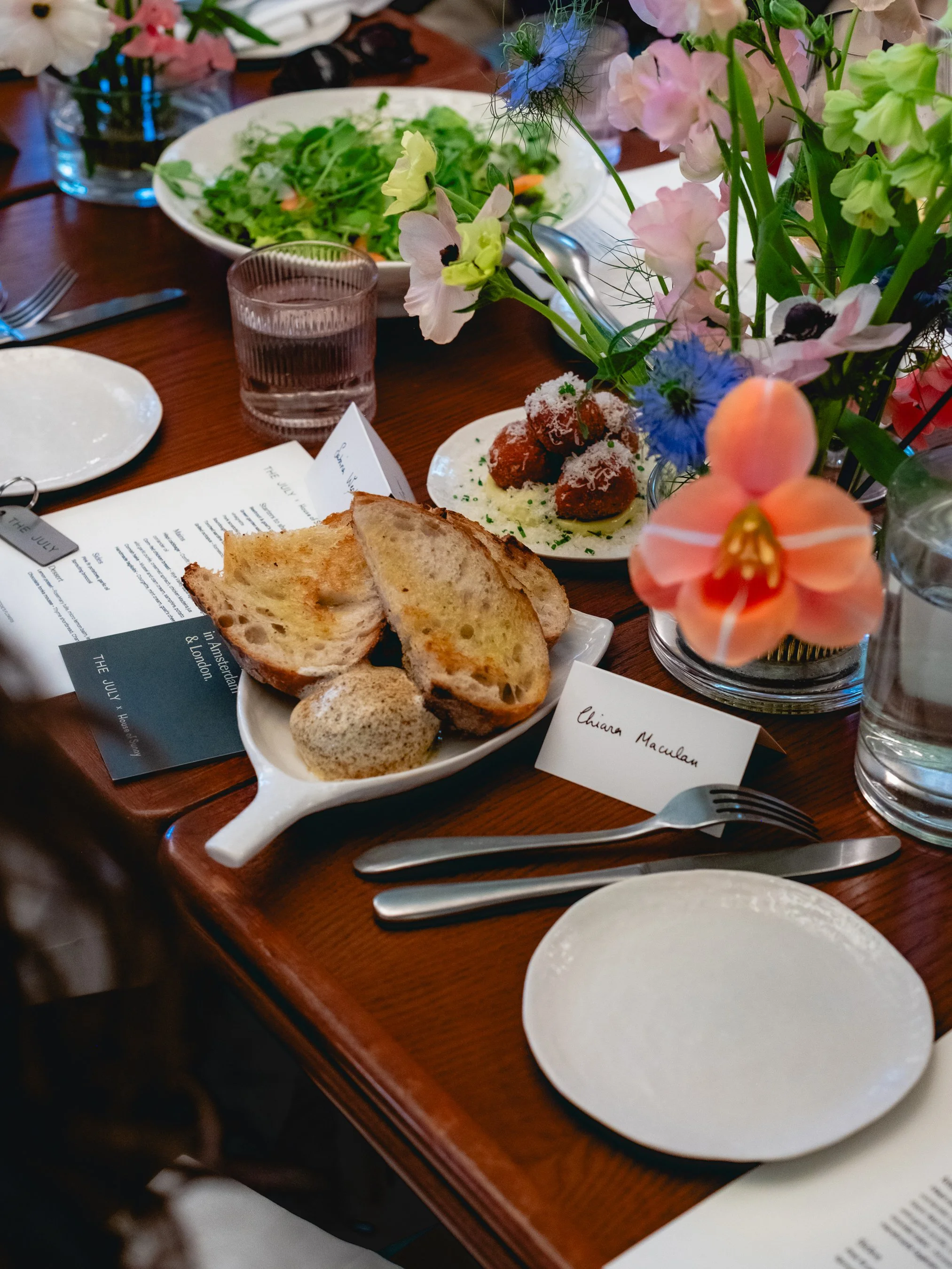 A table set with a variety of food including bread, salad, fried items on a plate, and soft drinks. There are flowers in vases as decoration, and place cards with names, including one labeled 'Chiara Maculan.' Silverware and empty plates are also vis