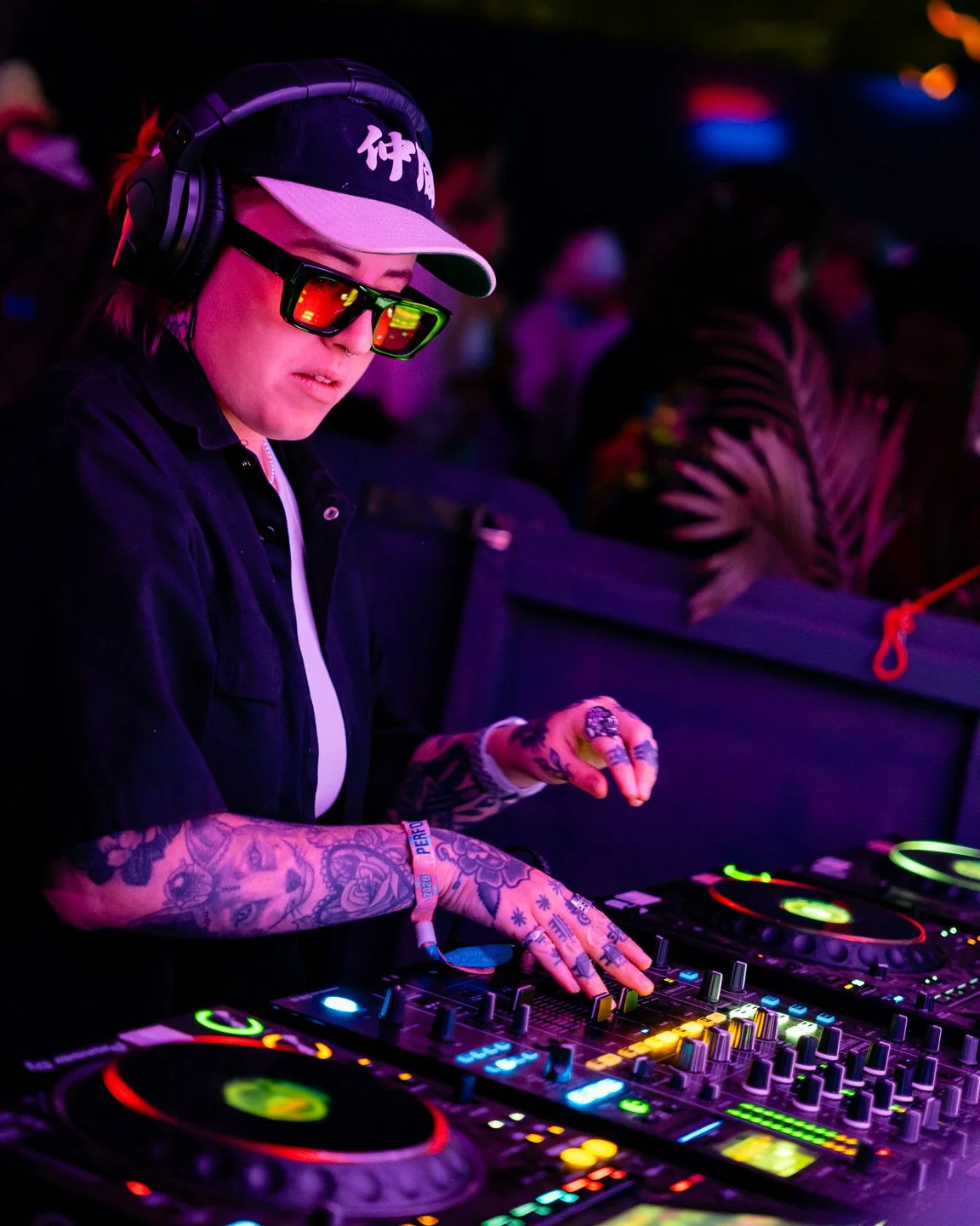 A female DJ wearing sunglasses, a cap, and headphones, operating a DJ mixing console at a dark, colorful music event.