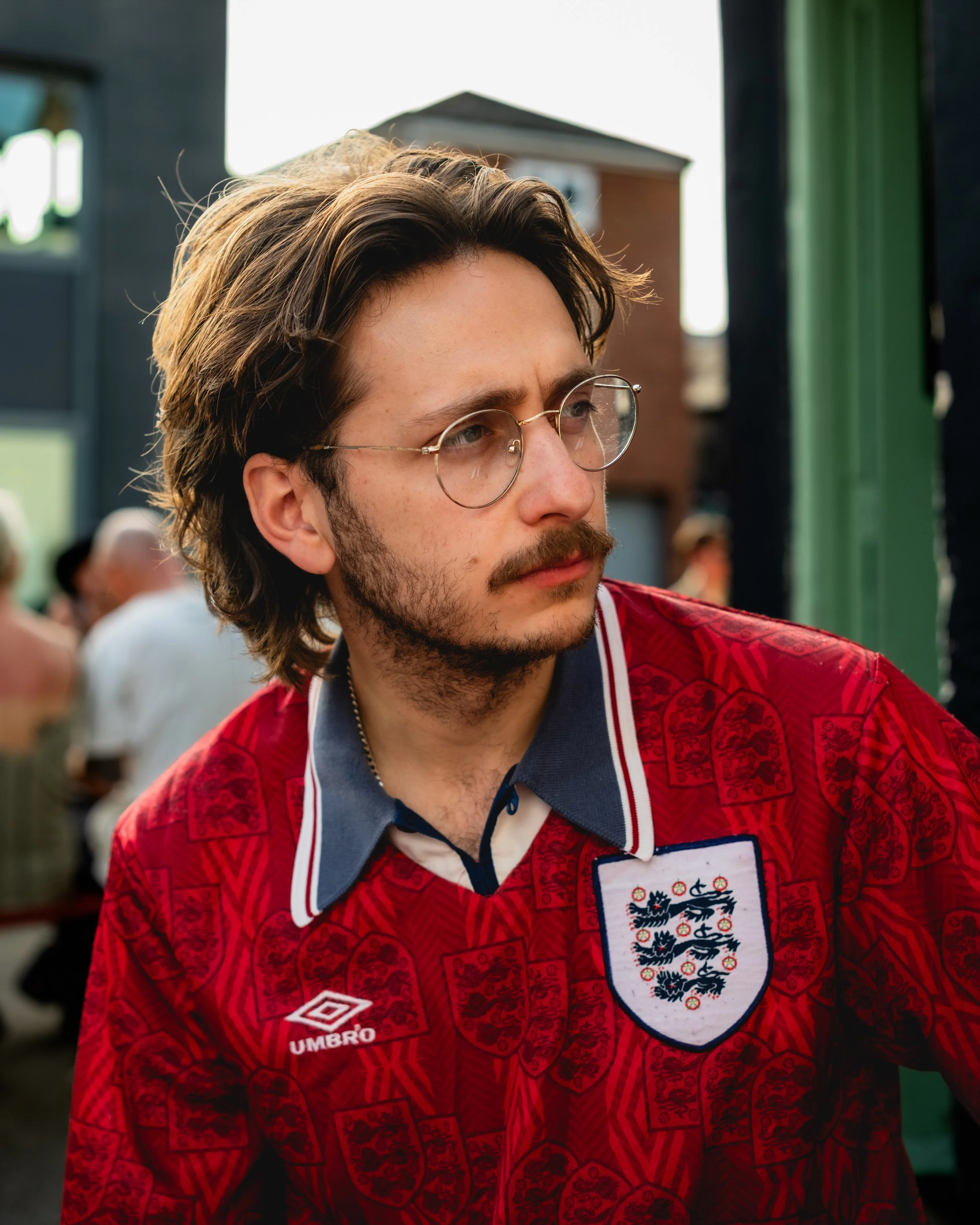 A man with long brown hair and glasses wearing a red England soccer jersey with the three lions crest, at an outdoor gathering.