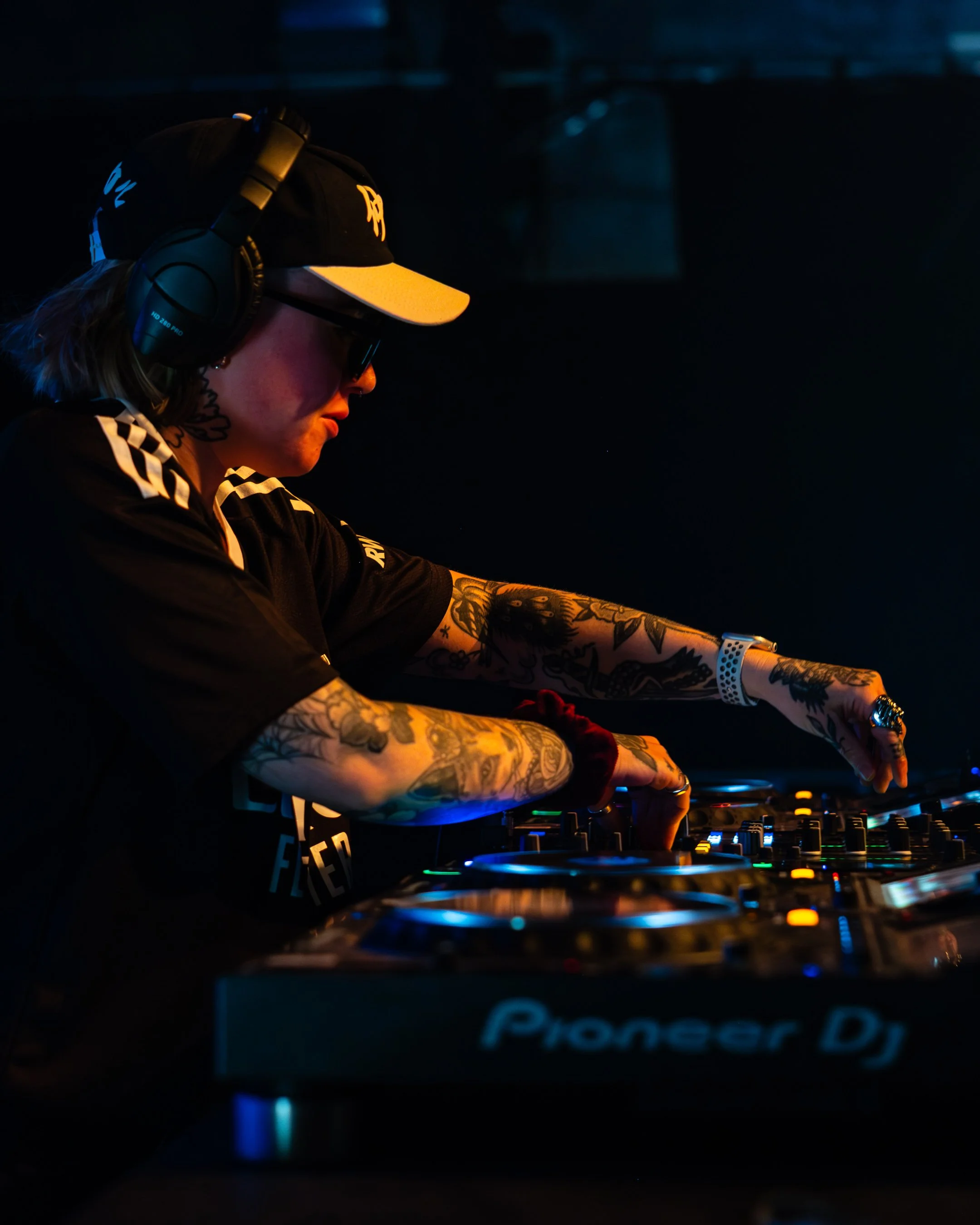 A DJ with tattoos on arms, wearing glasses, headphones, a black baseball cap, and a black shirt, performing at a dark venue with blue and yellow lighting.