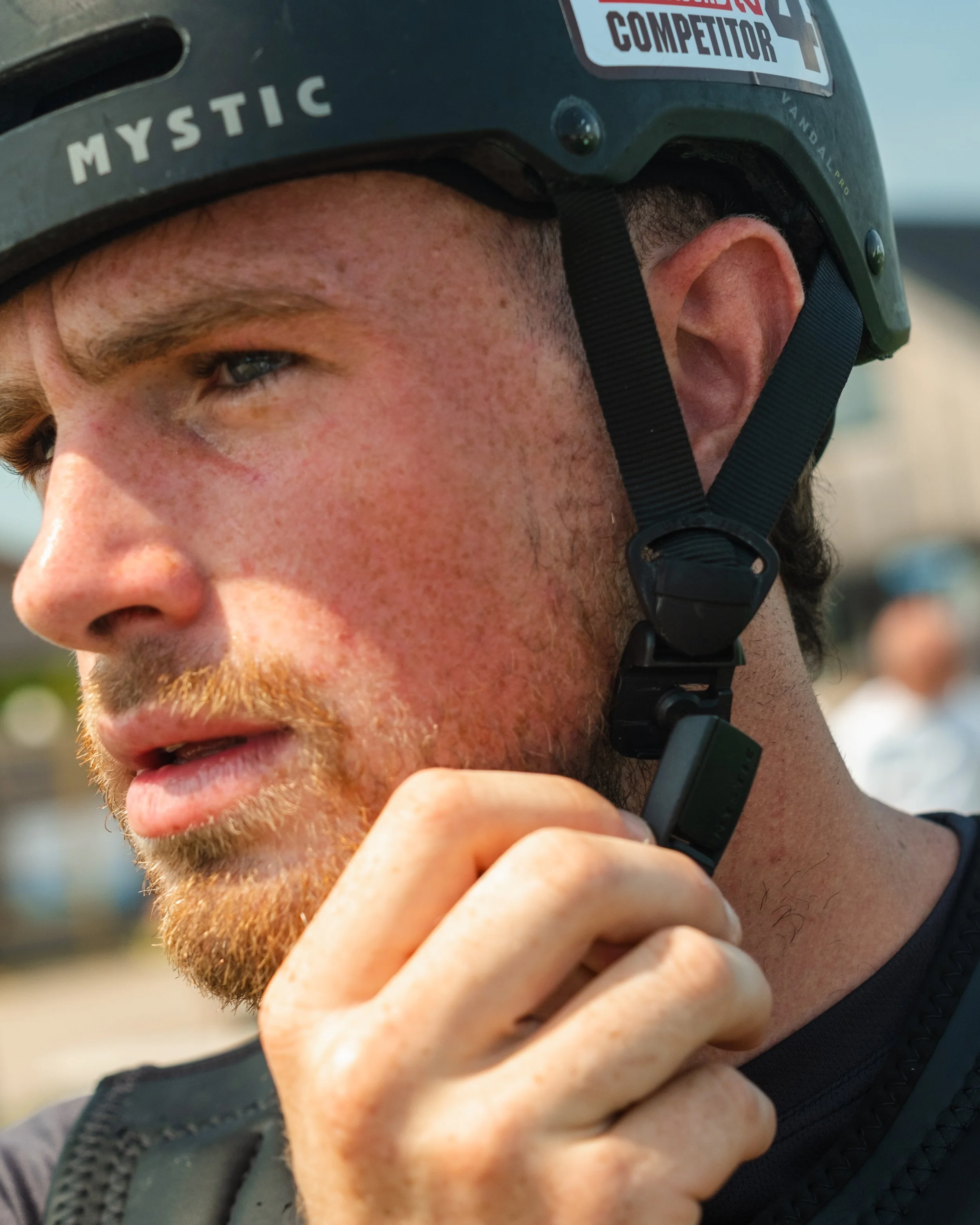 A close-up of a man wearing a black helmet with stickers, adjusting a microphone attached to his mouth. The helmet has stickers that say 'MYSTIC', 'COMPETITOR', and another sticker partially visible. The man has reddish hair, a beard, and is wearing 
