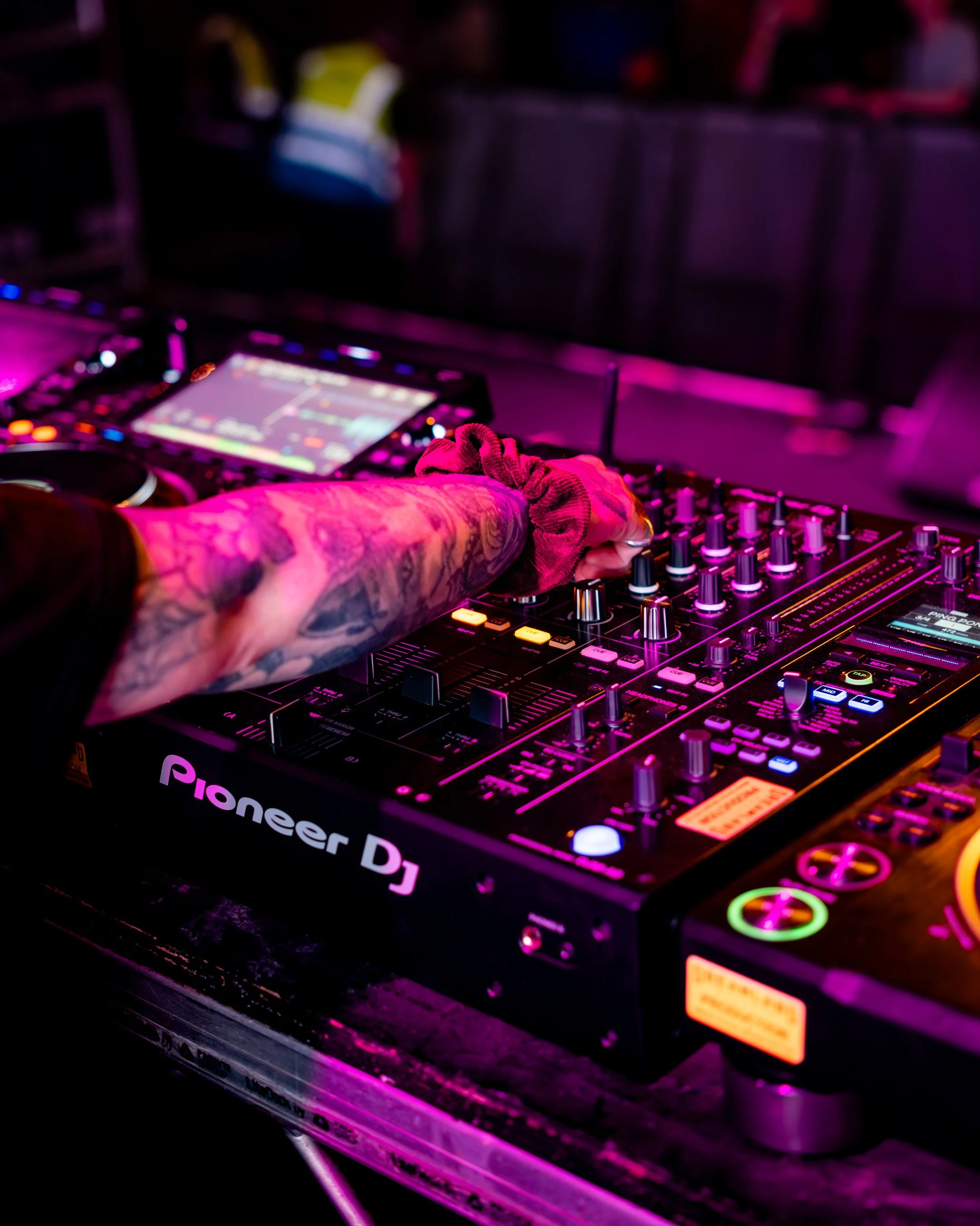 A DJ's hand with tattoos on arm adjusting controls on a Pioneer DJ mixer in a dark club with colorful LED lights.