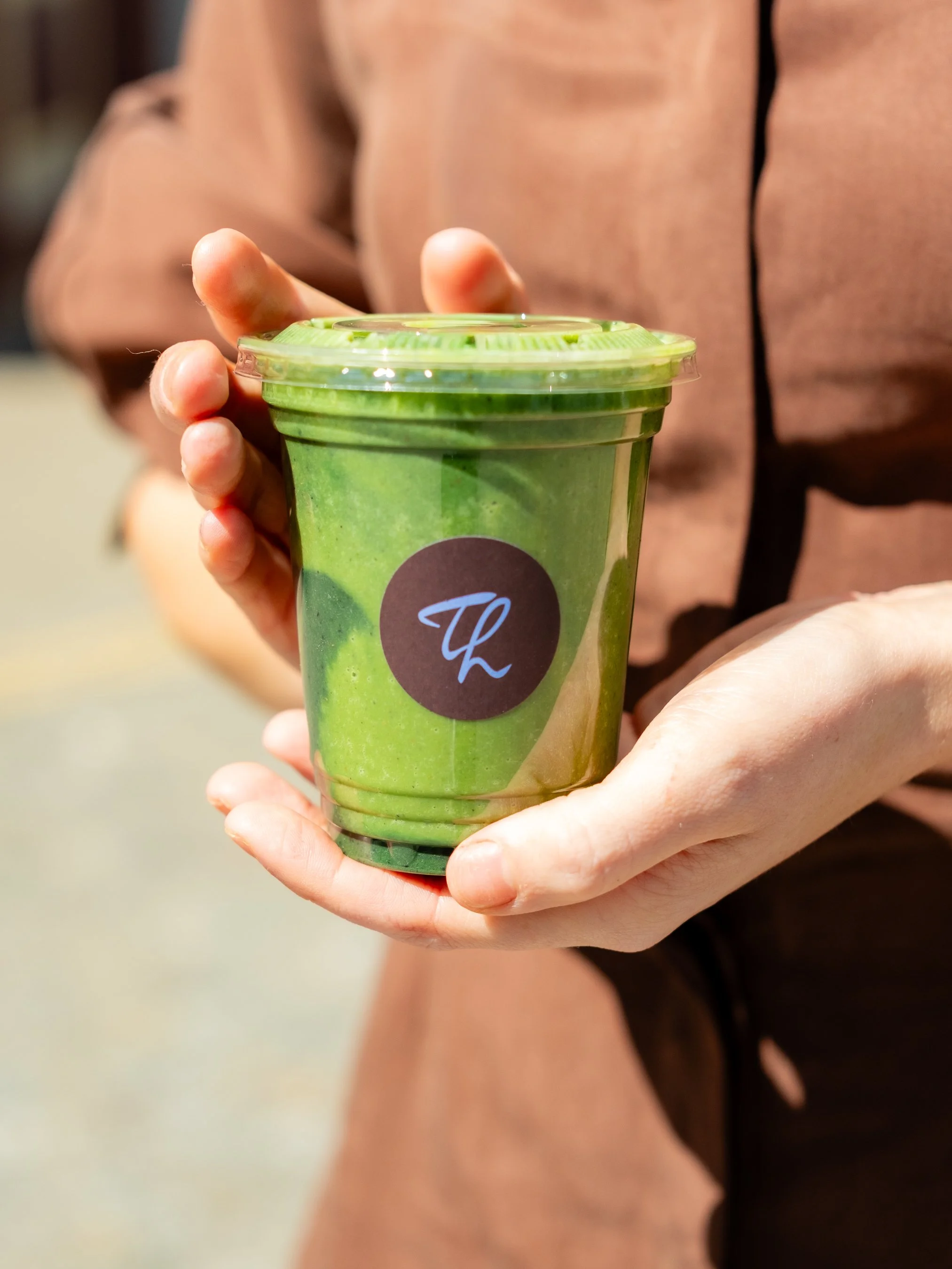 A person holding a green smoothie drink in a cup with a lid, featuring a black circular logo with a white 'T' and 'L' on it.