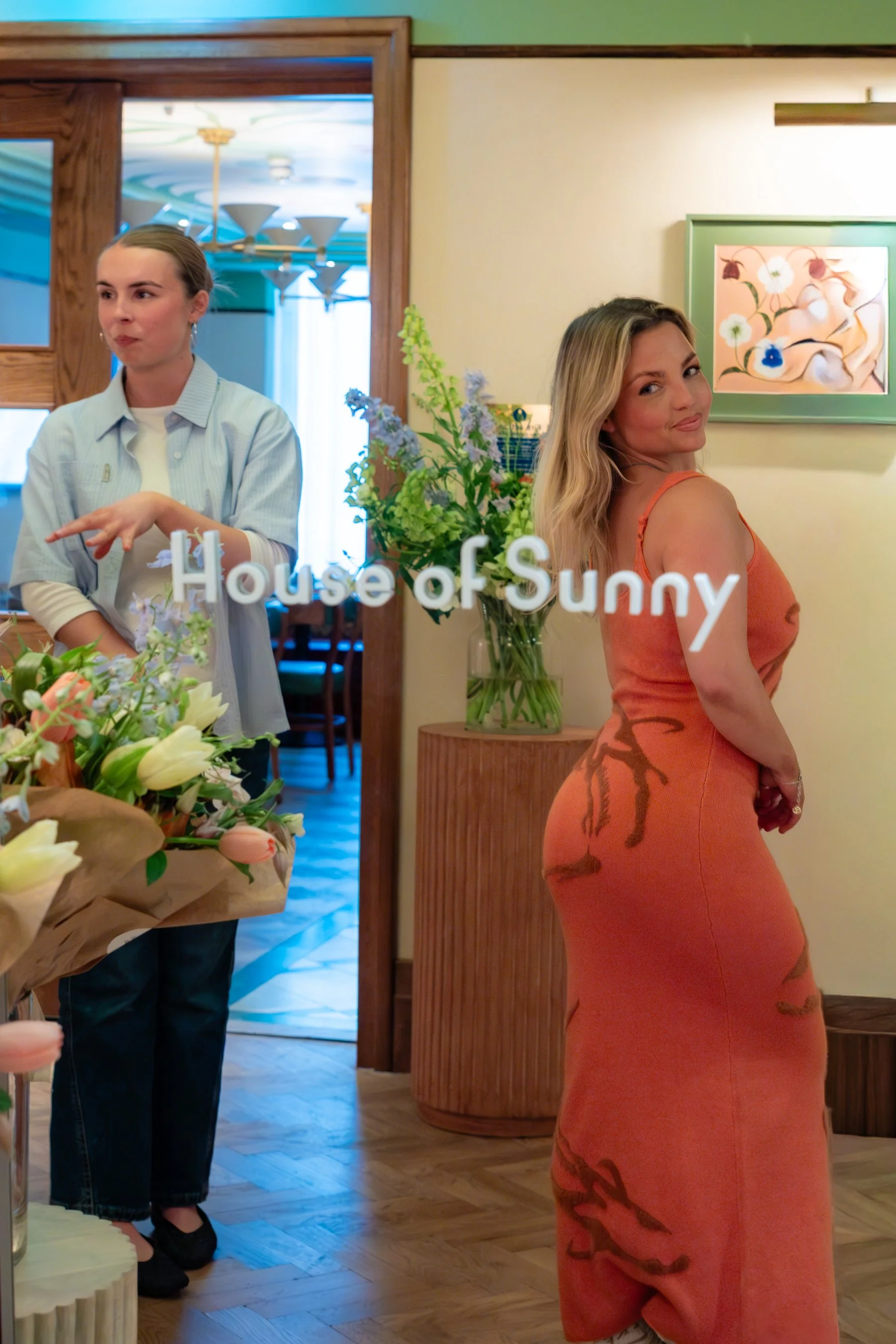 Two women standing inside a restaurant with tropical decor and floral artwork. One woman is a server wearing a light blue shirt, the other is a guest in an orange dress. There are flowers and wooden accents.