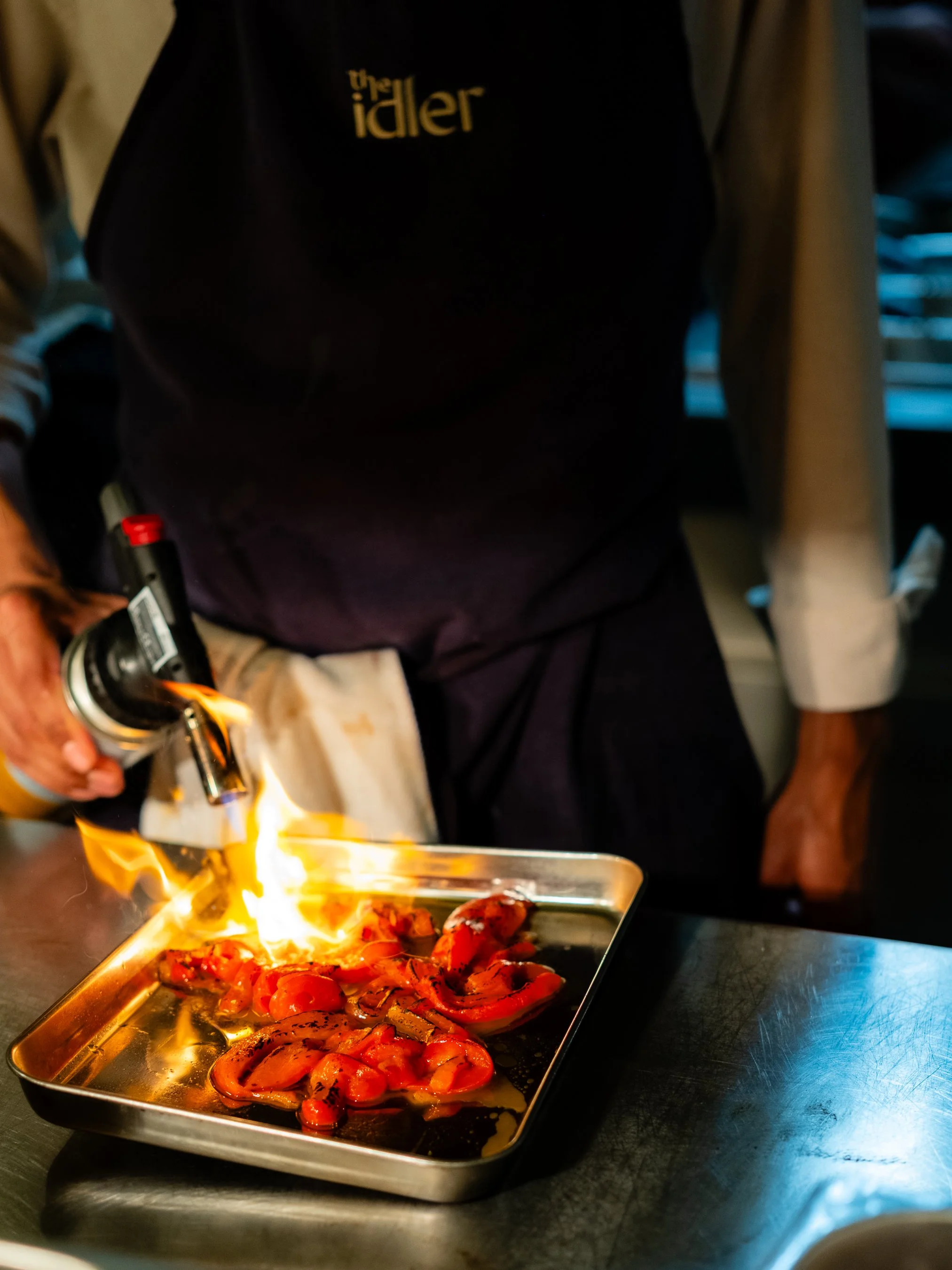 Chef igniting a flame with a blowtorch while preparing roasted tomatoes on a metal tray in a professional kitchen.