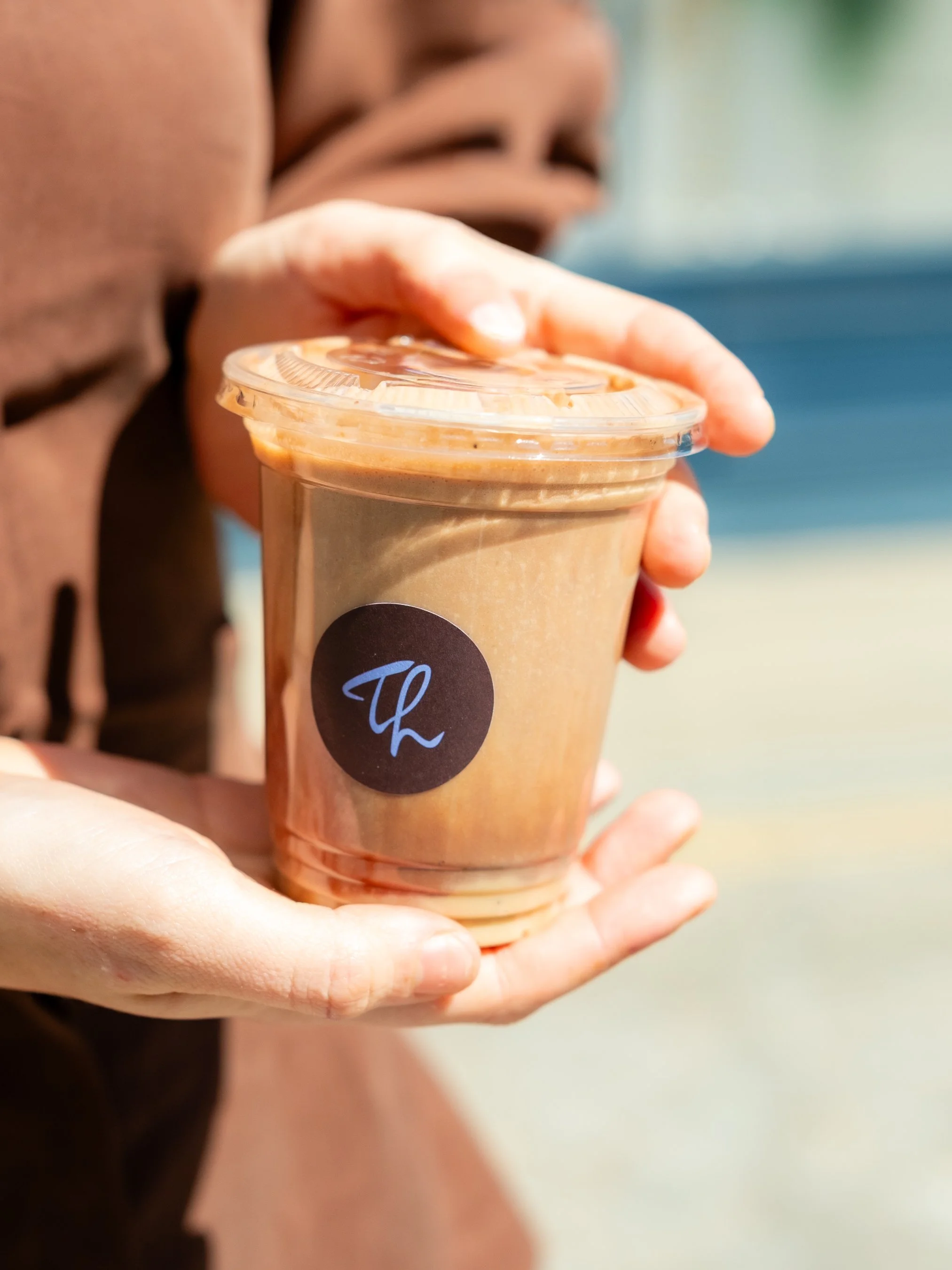 A person holding a plastic cup filled with a brown iced coffee or latte, with a black circular label showing a white cursive letter 'L'.