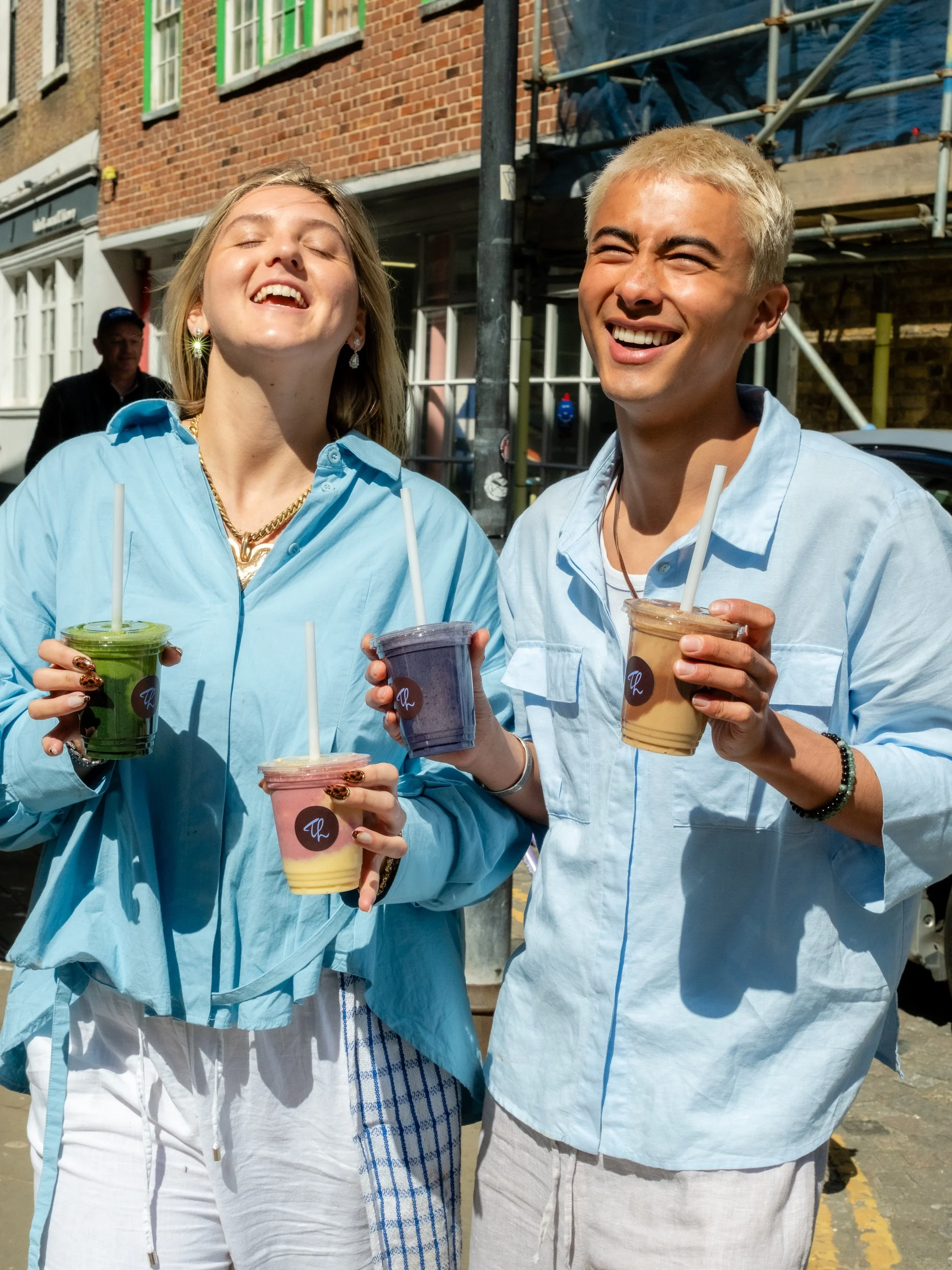 Two smiling young adults, a woman and a man, holding colorful drinks with straws outdoors on a sunny day, with a brick building and scaffolding in the background.
