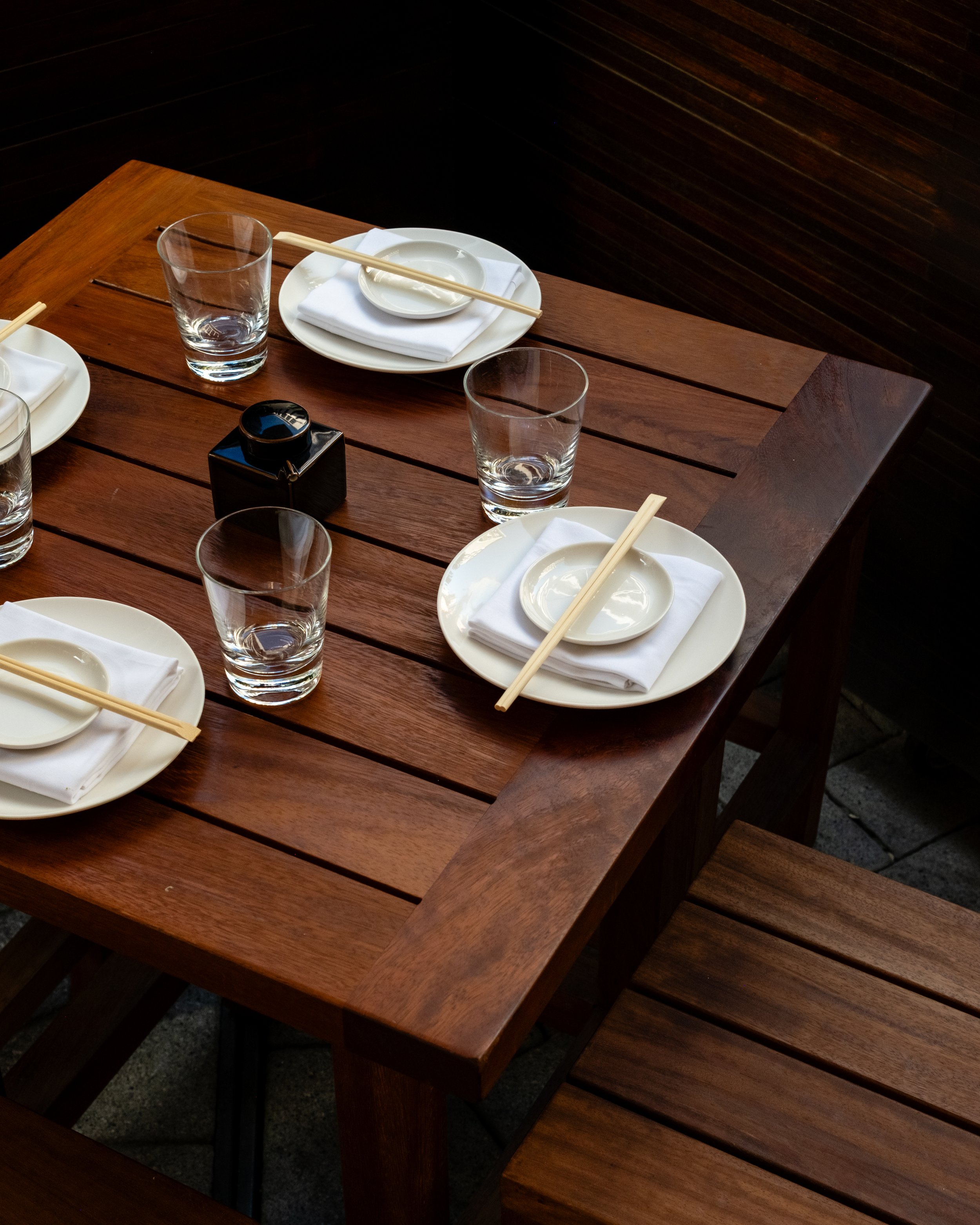 A wooden outdoor table set with white plates, napkins, chopsticks, empty small bowls, and glasses, with a dark wooden wall in the background.