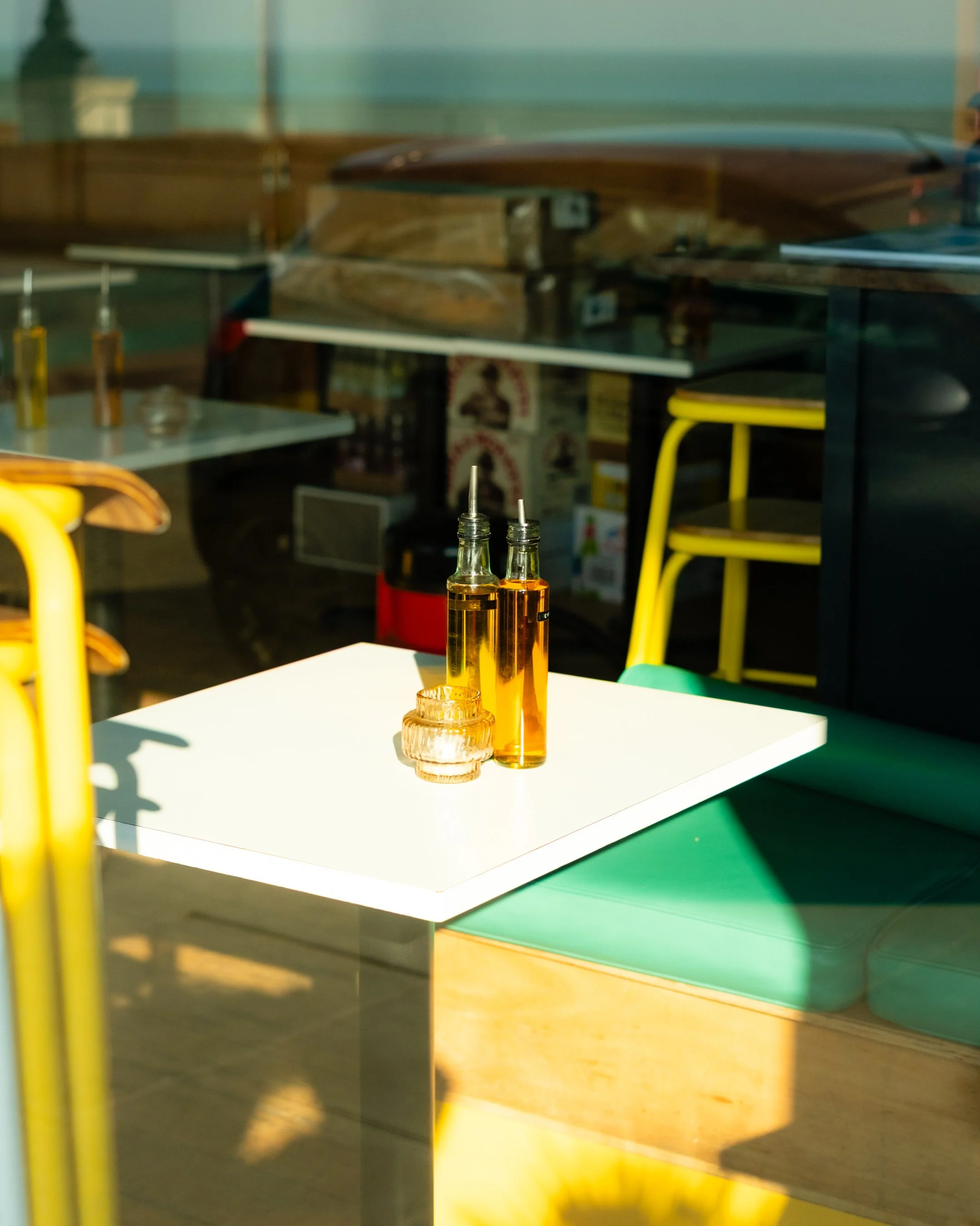 Reflection of a dining area with yellow chairs, a white table, and a few condiment bottles, seen through a glass window.