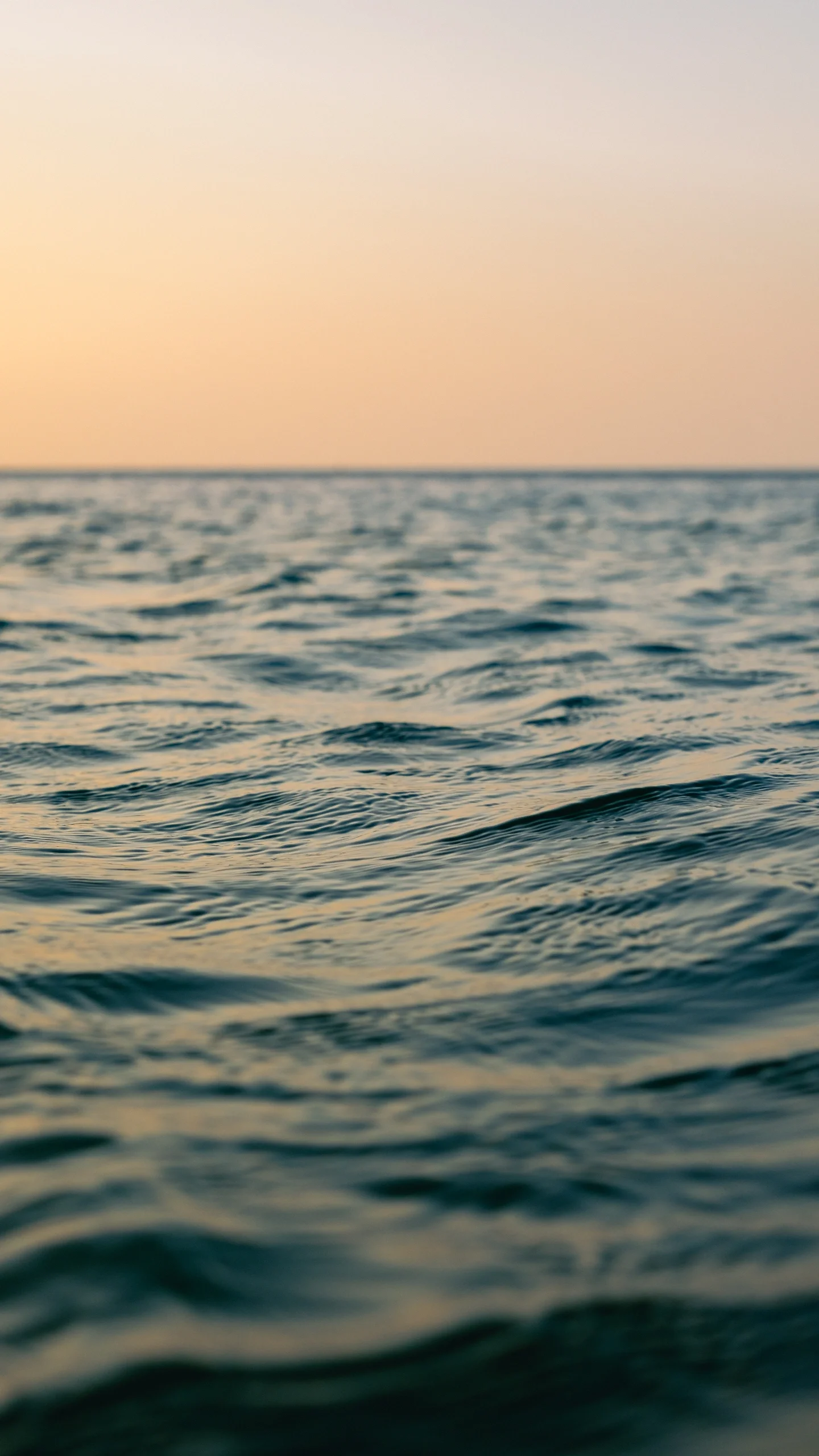 A calm ocean surface with gentle ripples, under a pastel-colored sky at sunset or sunrise.