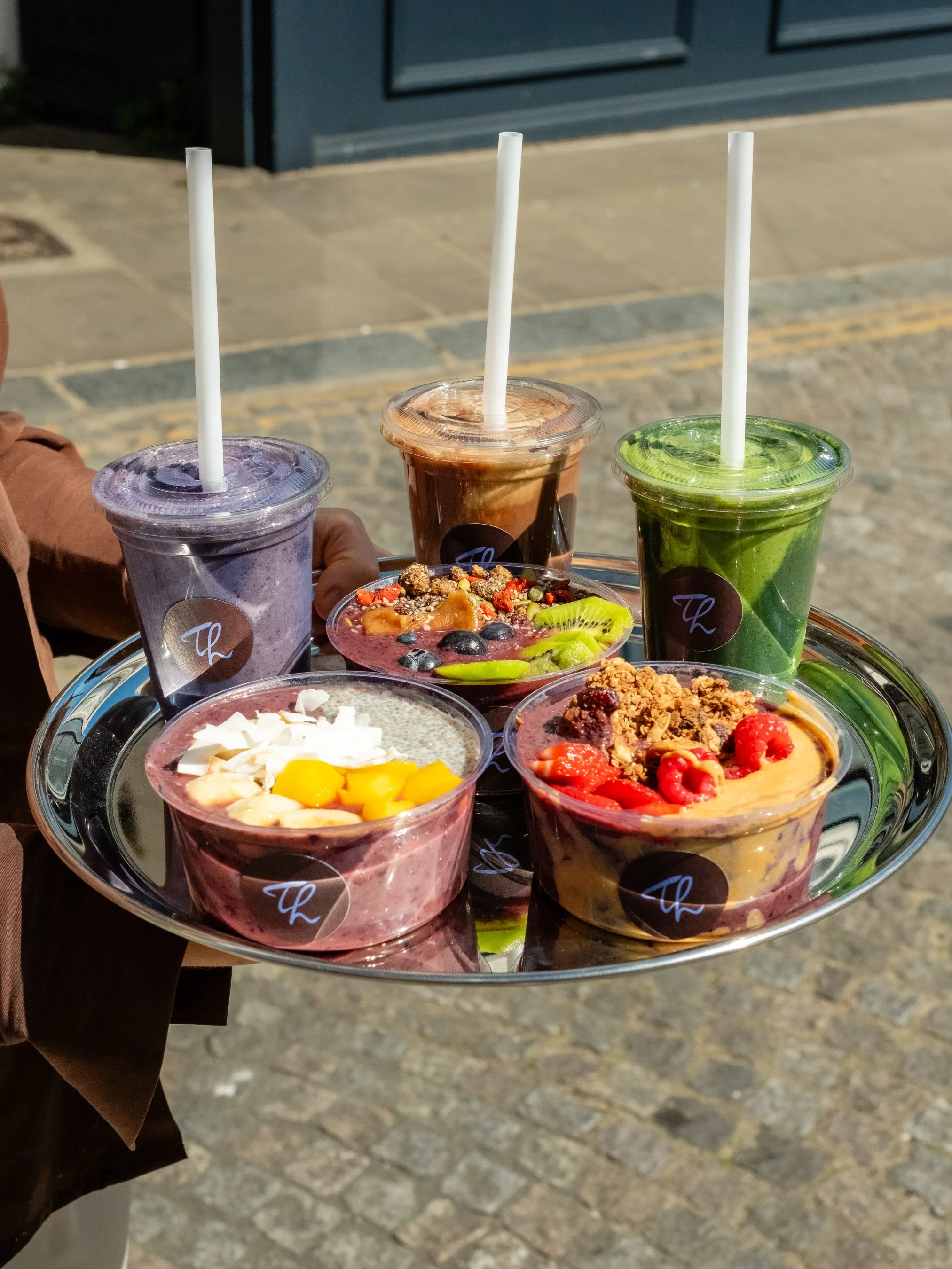 Tray of assorted frozen yogurt or smoothie cups in various flavors, with toppings like fruit, granola, and syrup, outside on a sunny day.