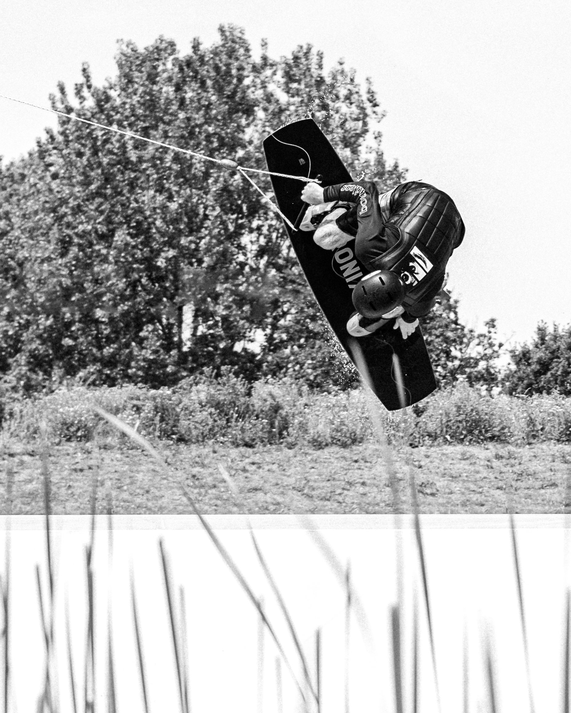 A skateboarder, wearing a helmet and protective gear, performs an aerial trick in the air against a backdrop of trees and a partly cloudy sky.