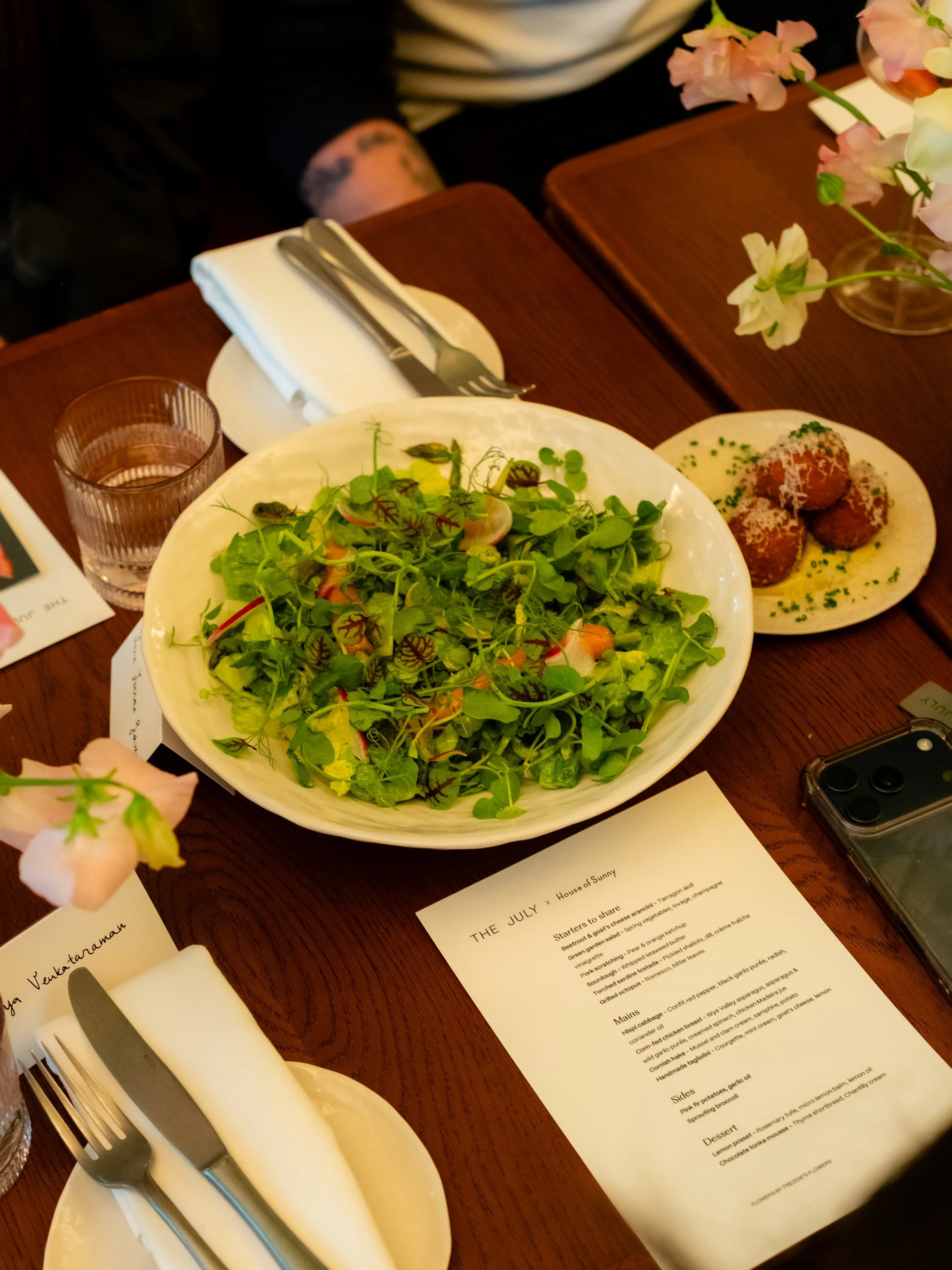 A table set with a large bowl of mixed greens salad, a plate of croquettes with dipping sauce, a glass of water, a knife and fork on a folded napkin, a flower arrangement, and a menu.