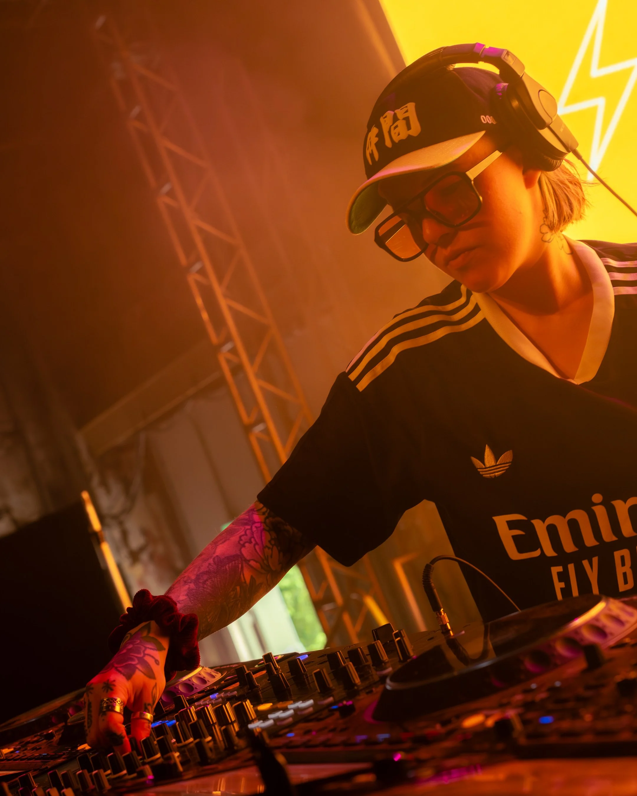 A DJ wearing a black Adidas shirt, a watch, a beige cap, and large glasses, with tattoos on their arm, operating DJ equipment in a vibrant, colorful setting with yellow and orange lighting.